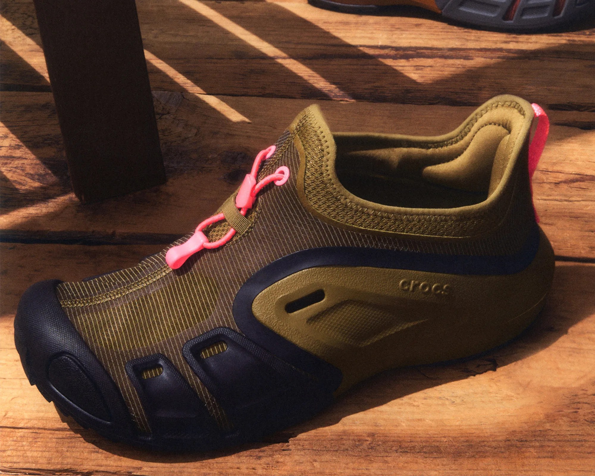 Olive green and black Crocs shoe with pink laces on a wooden surface.