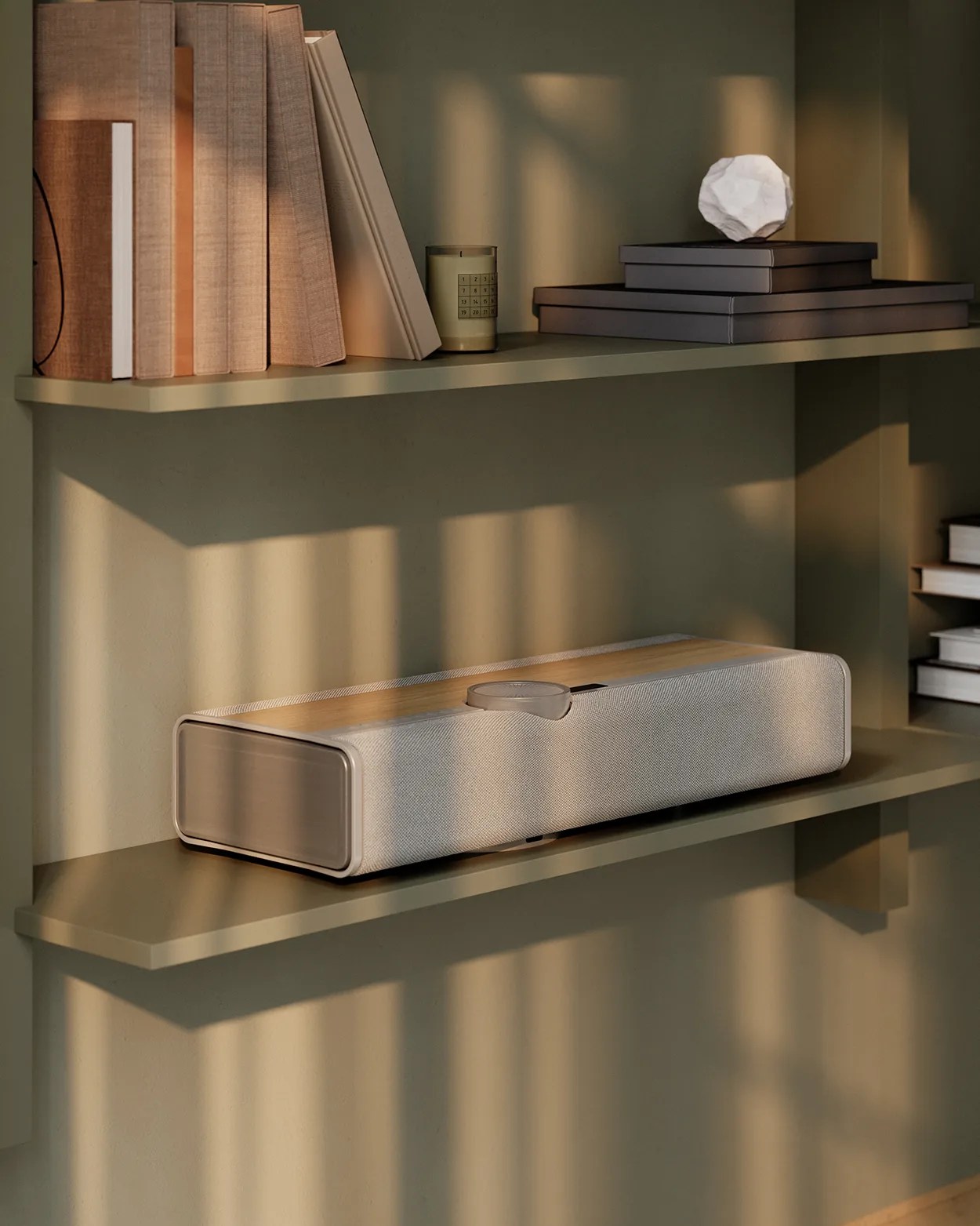 Rectangular beige and wood-finished speaker on an olive green wall shelf with books and decorative items.