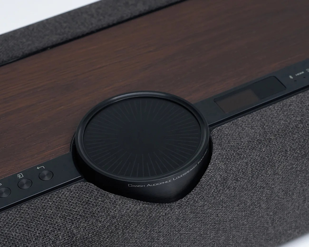 Close-up of a black circular control dial on a dark wood and gray fabric speaker panel with buttons.