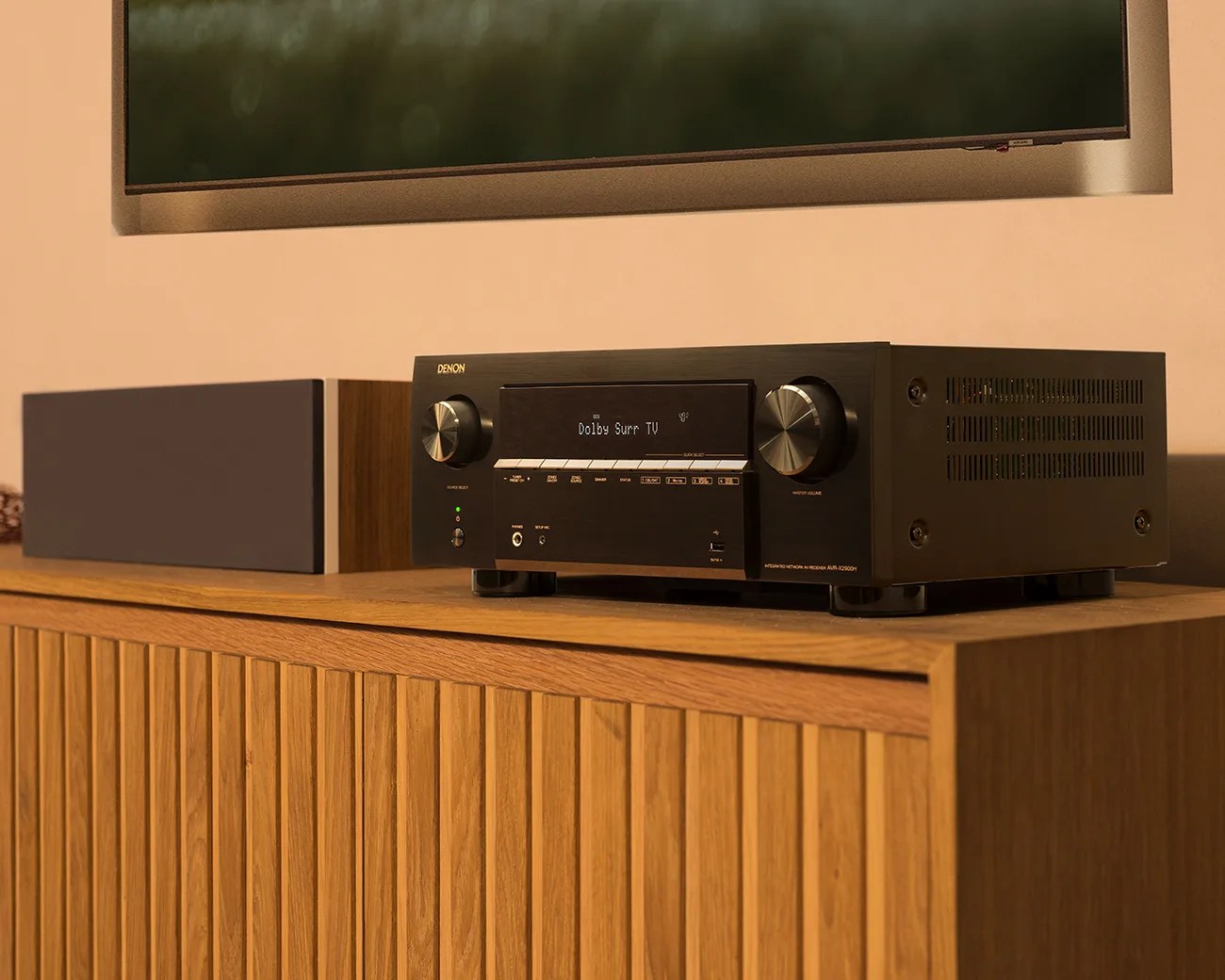 Black Denon AV receiver with silver knobs and digital display on a wooden cabinet next to a center speaker.
