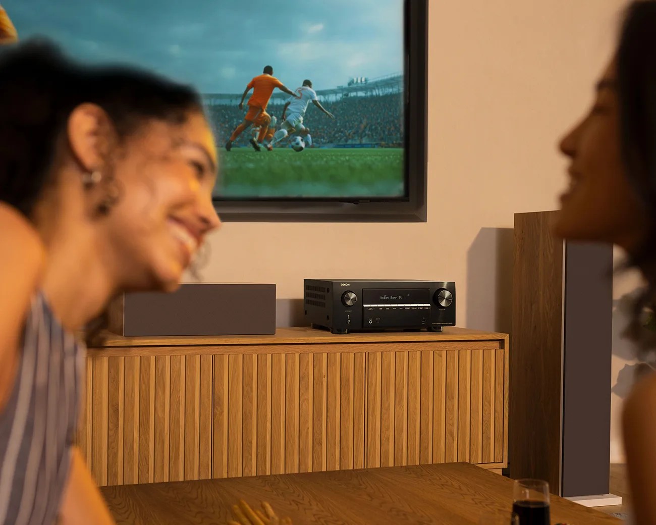 Black Denon AV receiver and brown speaker on a wooden cabinet under a wall-mounted TV showing a soccer game.