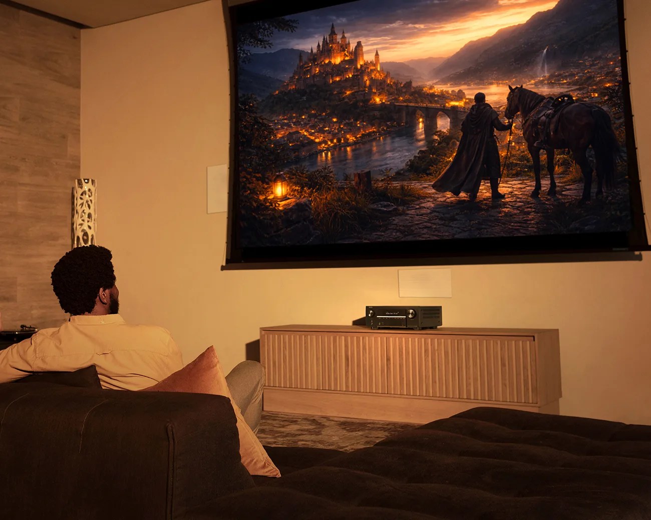 Man sitting on a dark brown sofa watching a large wall-mounted screen showing a fantasy castle scene.