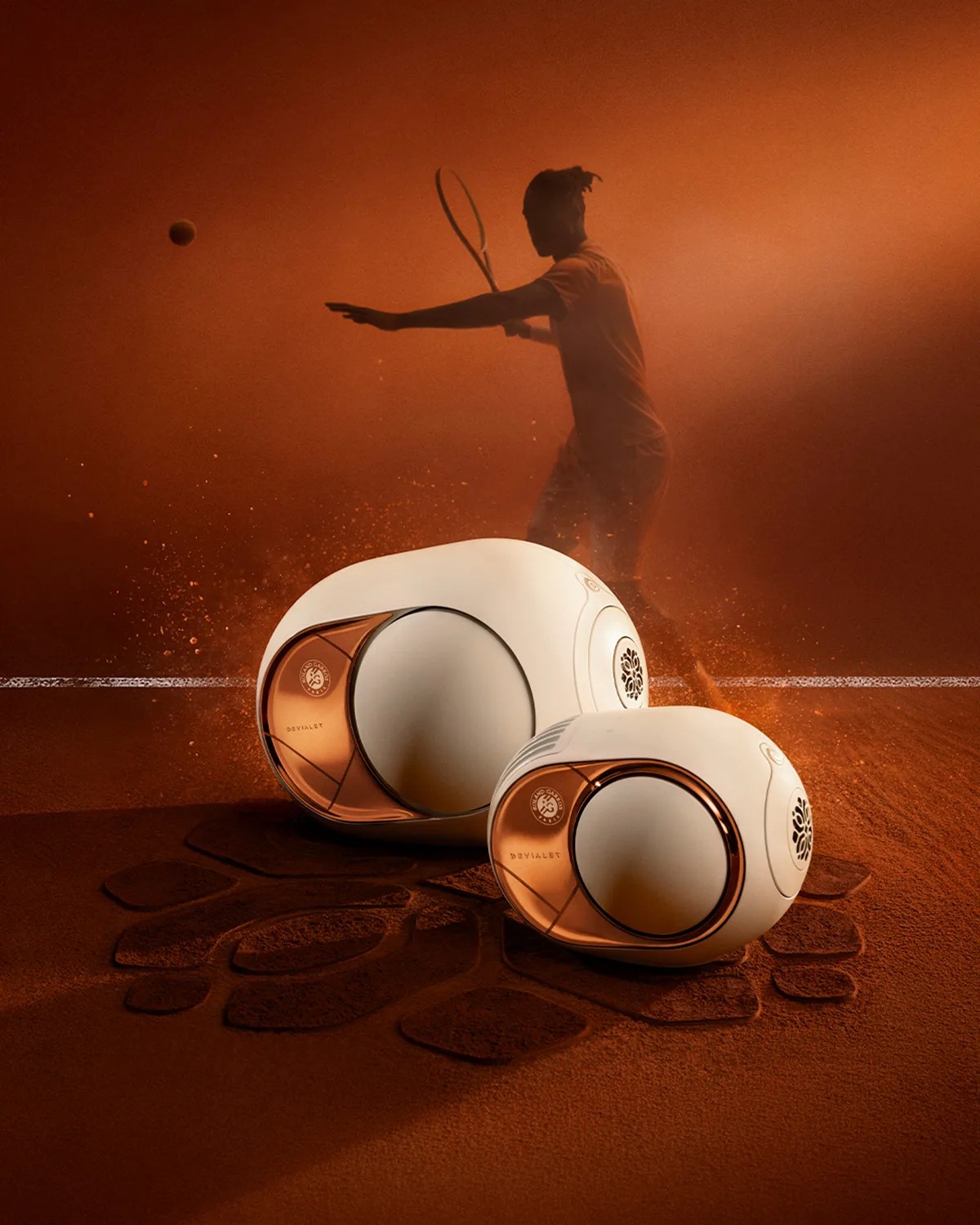 Two white and copper Devialet speakers on a clay tennis court with a silhouette of a tennis player in the background.