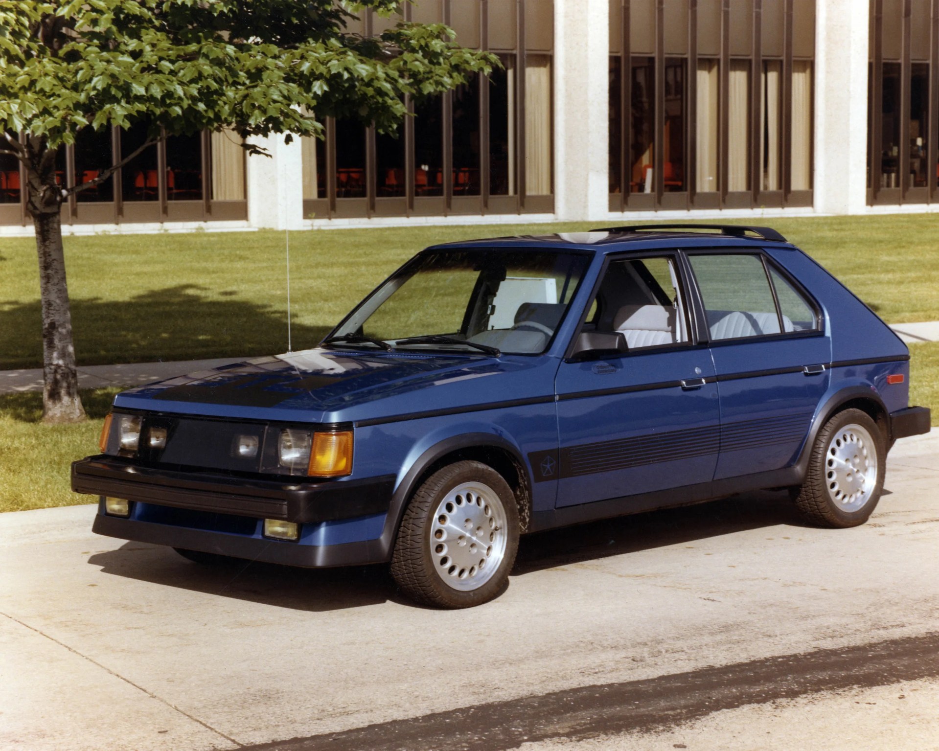 Blue 1980s hatchback car parked on a driveway near a tree and a building with large windows.