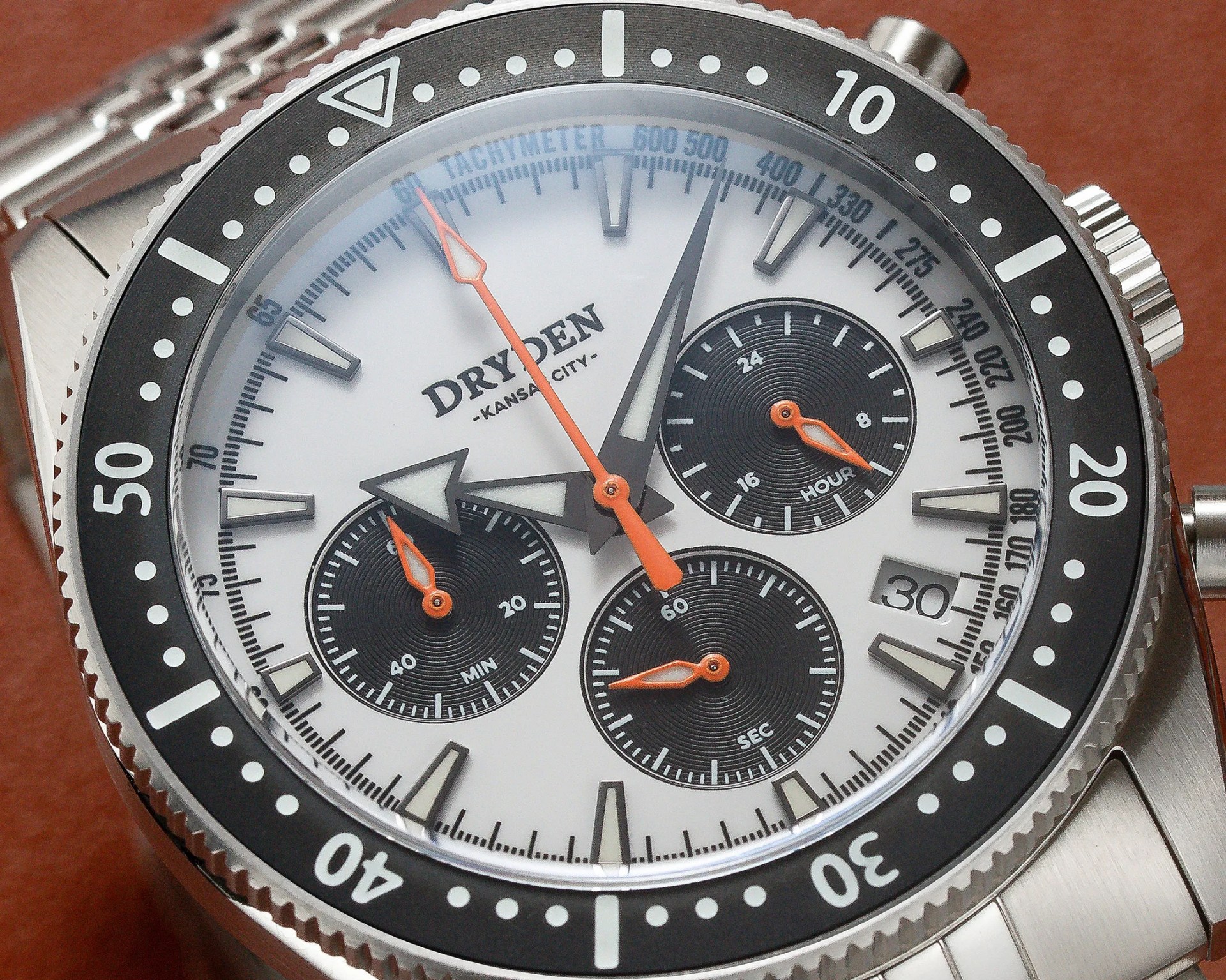 Close-up of a Dryden Kansas City chronograph watch with white dial, black subdials, orange hands, and stainless steel bezel and bracelet.