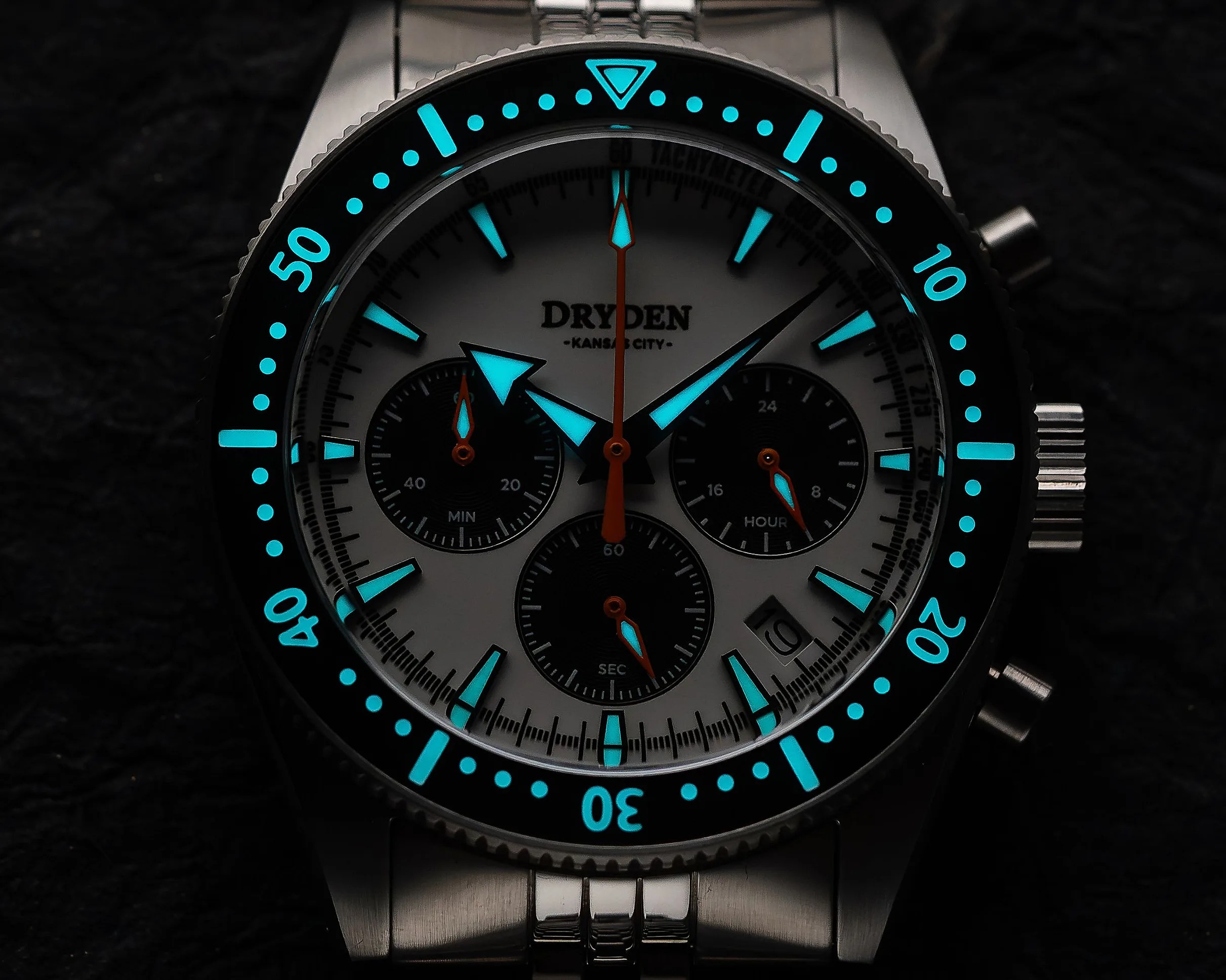 Close-up of a Dryden Kansas City wristwatch with glowing blue hour markers and hands on a silver metal band.