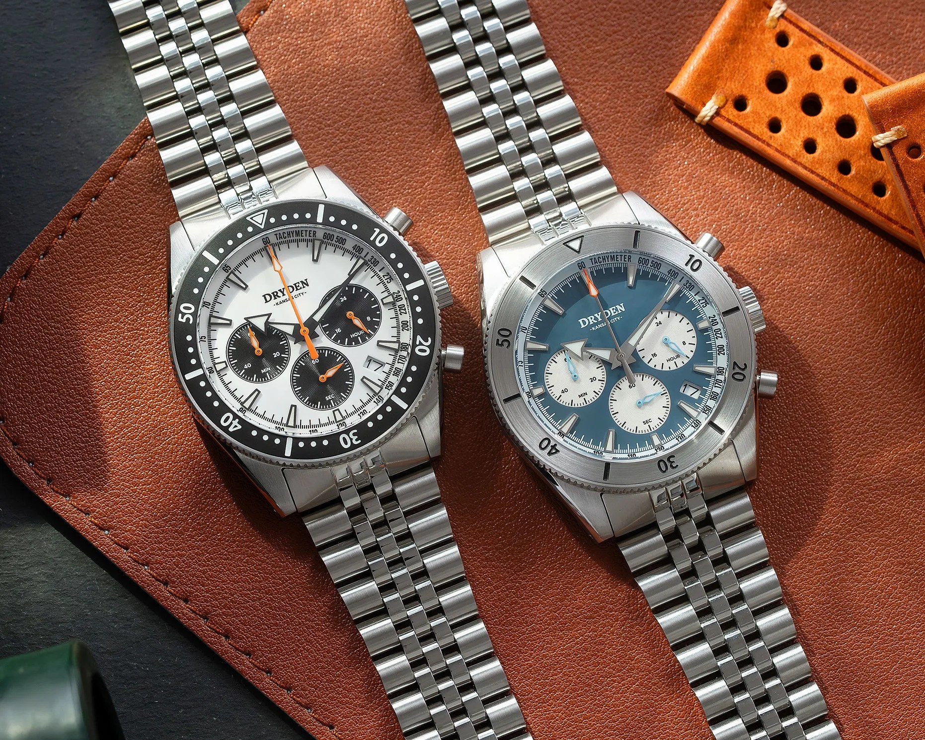 Two Dryden chronograph watches with stainless steel bracelets, one with a white dial and black bezel, the other with a blue dial and silver bezel, on a brown leather surface.