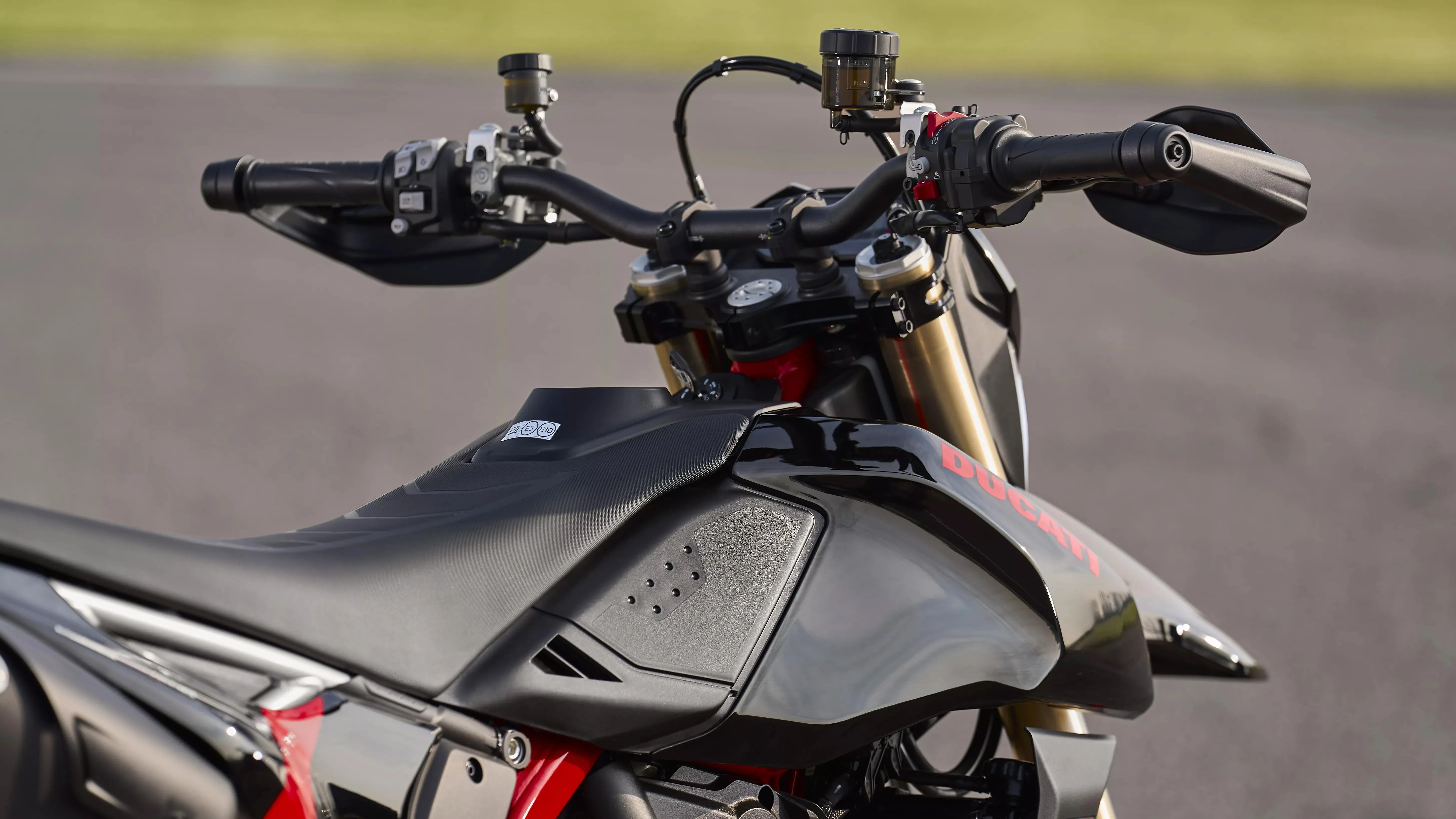 Close-up of black Ducati motorcycle fuel tank and handlebars with control switches and handguards.