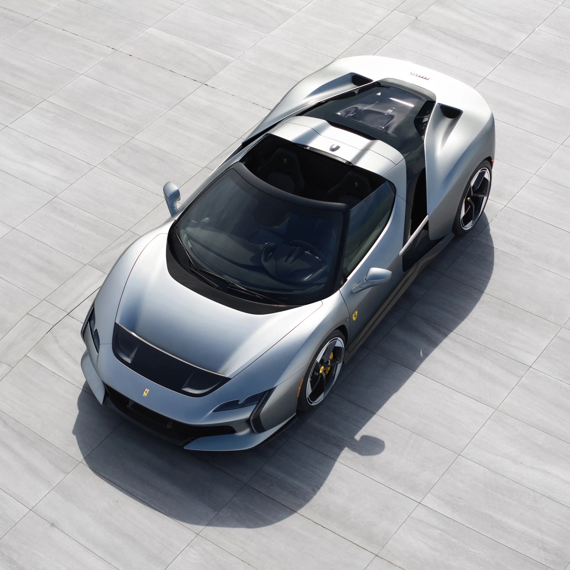 Silver Ferrari sports car with black accents parked on light gray tiled ground viewed from above.