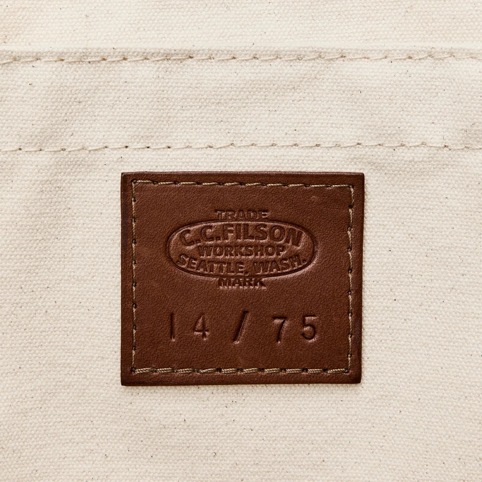 Brown leather patch with embossed text "C.C. Filson Workshop Seattle, Wash. Mark" and "14/75" on beige canvas fabric.