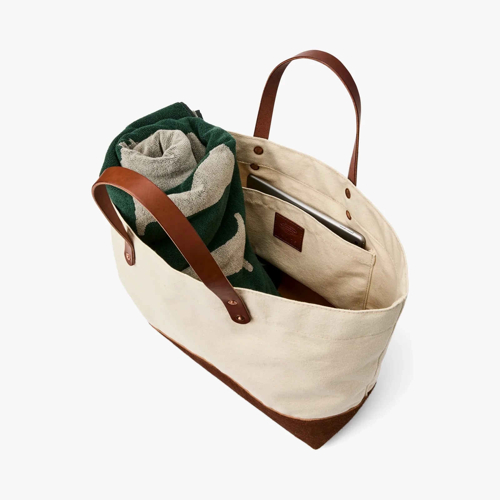 Beige canvas tote bag with brown leather handles and bottom, containing a rolled green and beige towel and a tablet in an inner pocket.