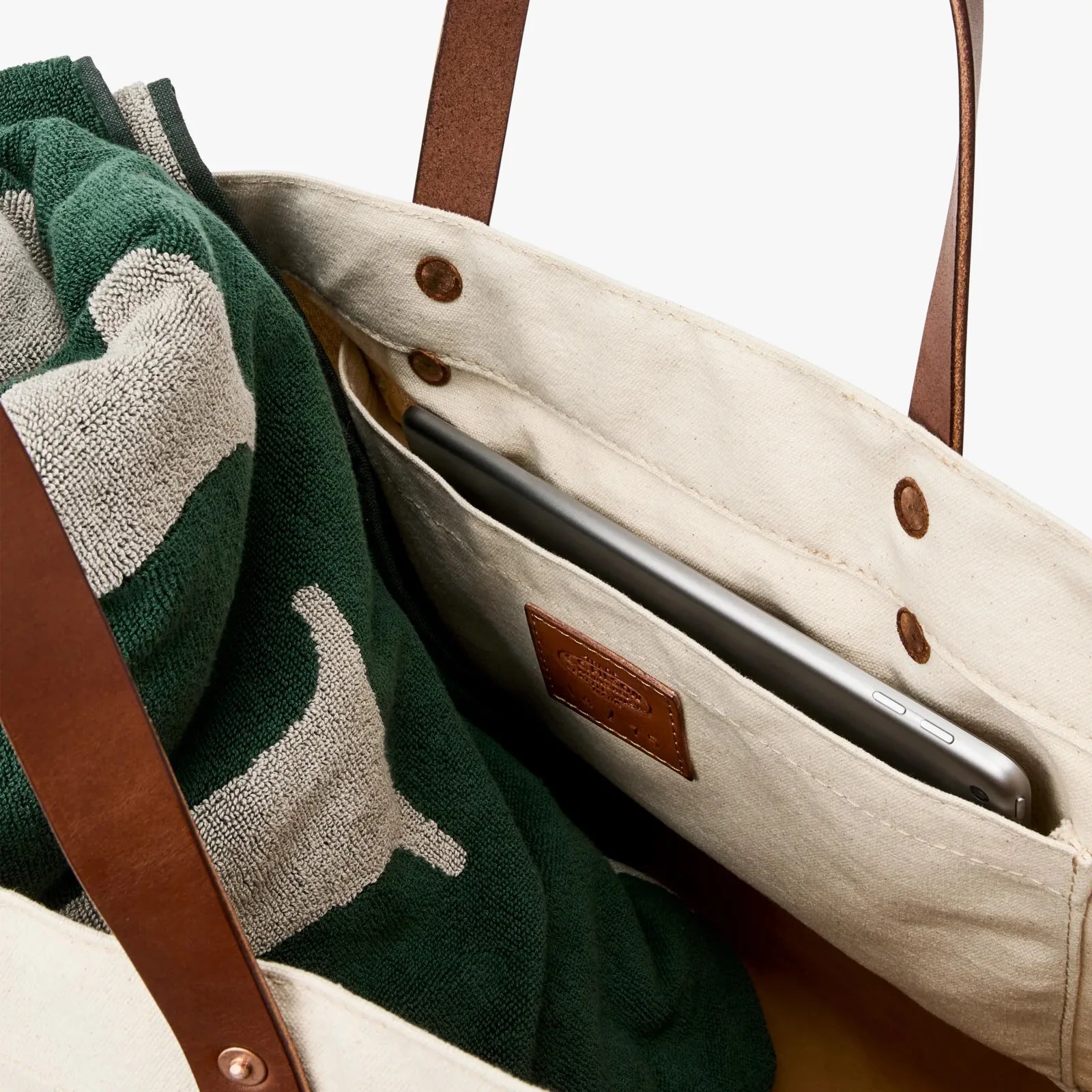 Beige canvas tote bag with brown leather handles, containing a green and beige towel and a silver tablet in an inner pocket.