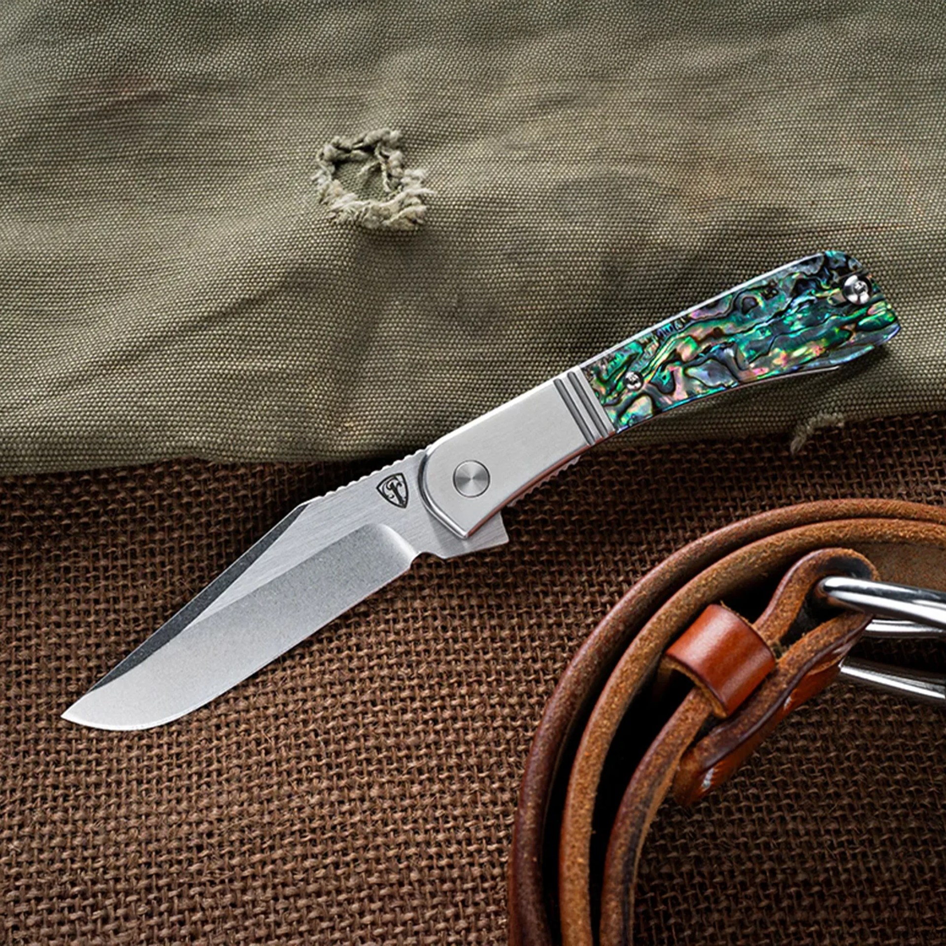 Folding knife with a silver blade and iridescent abalone handle resting on textured brown and green fabric next to a coiled leather strap.