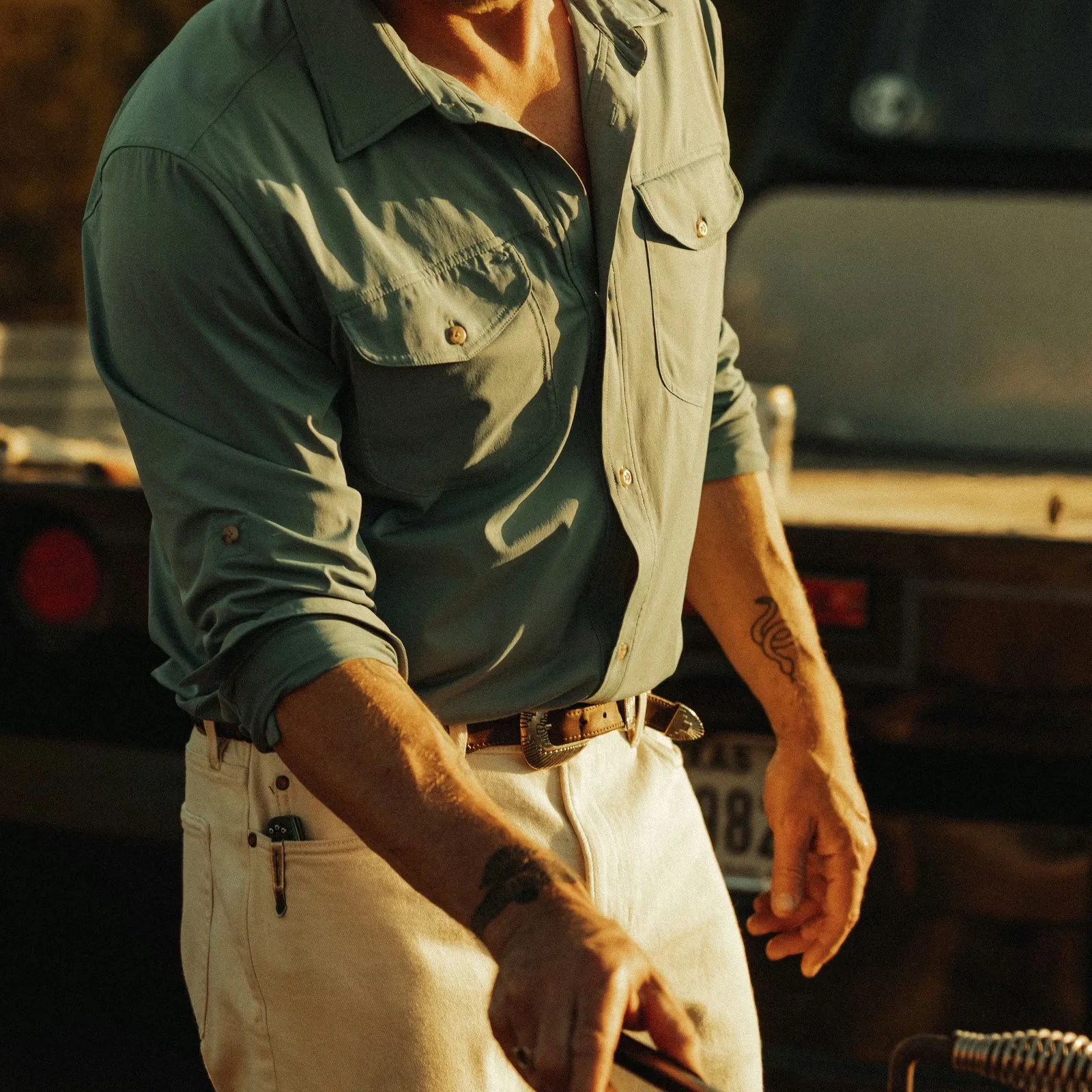 Olive green button-up shirt with chest pockets and rolled-up sleeves worn with white pants and a brown belt.