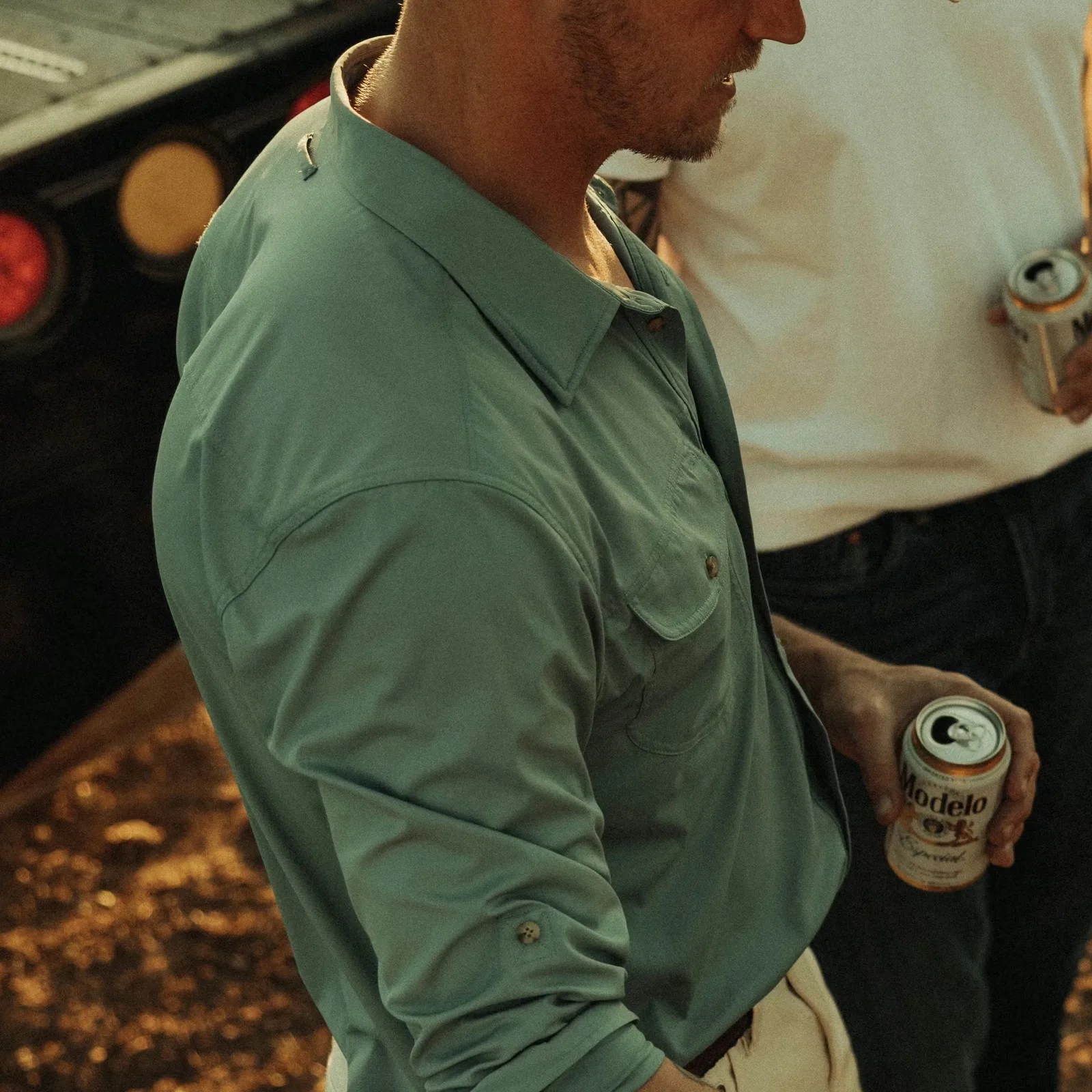 Man wearing a teal button-up shirt holding a can of Modelo beer with another person in a white shirt in the background.