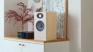 Beige and wood-finish bookshelf speaker on a wooden cabinet next to a black audio device and a vase with red branches.