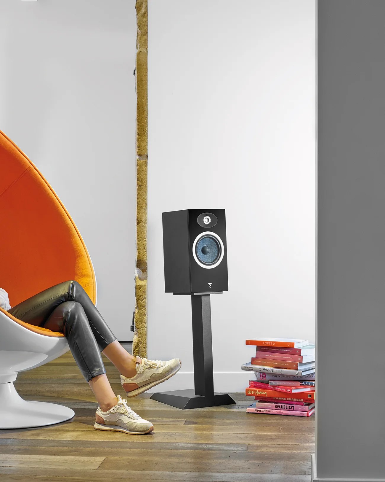 Black speaker with blue cone on a black stand next to a stack of books and an orange chair with a person sitting.