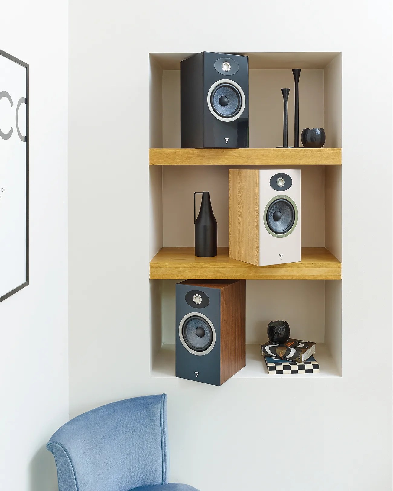 Three bookshelf speakers in black, white, and blue wood finishes on built-in wooden shelves with black decorative vases and stacked books nearby.