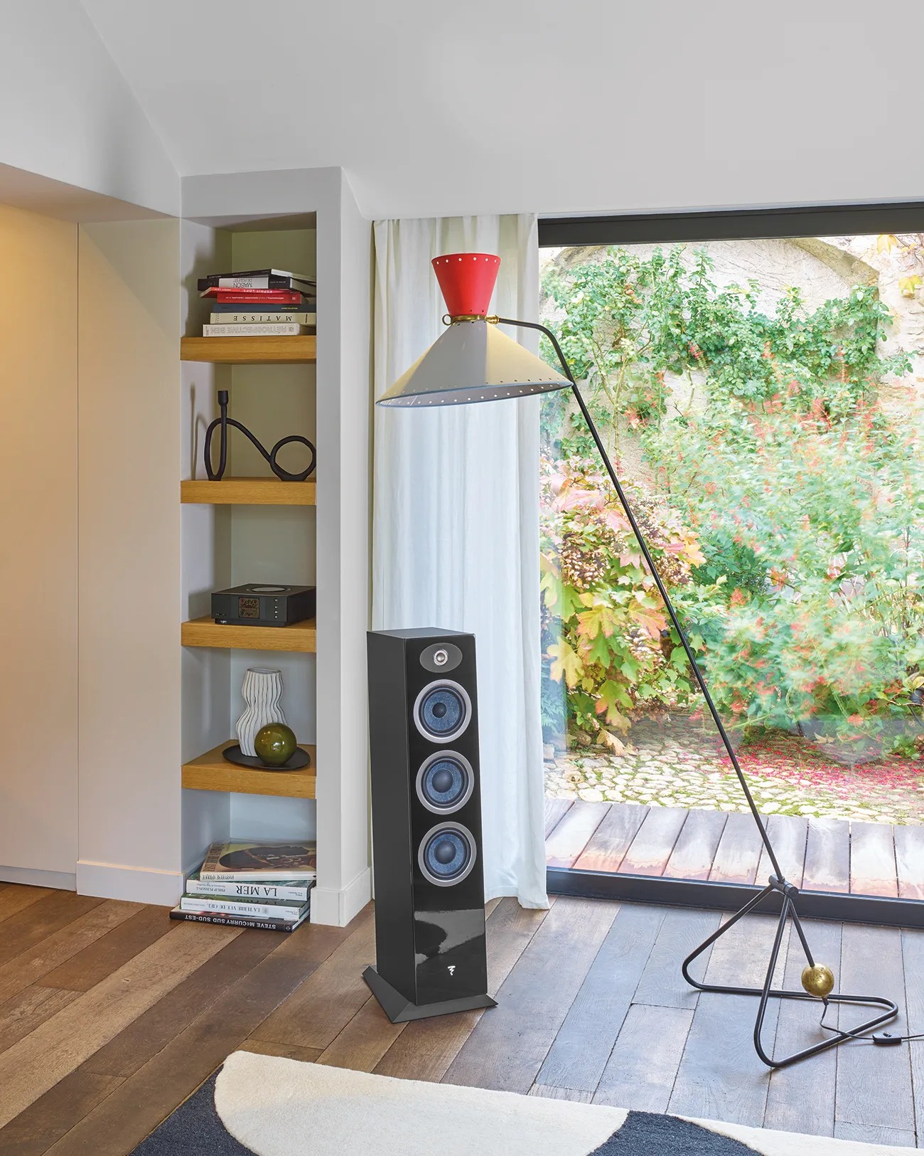 Black floor-standing speaker with three blue drivers next to a modern floor lamp and wooden shelves.