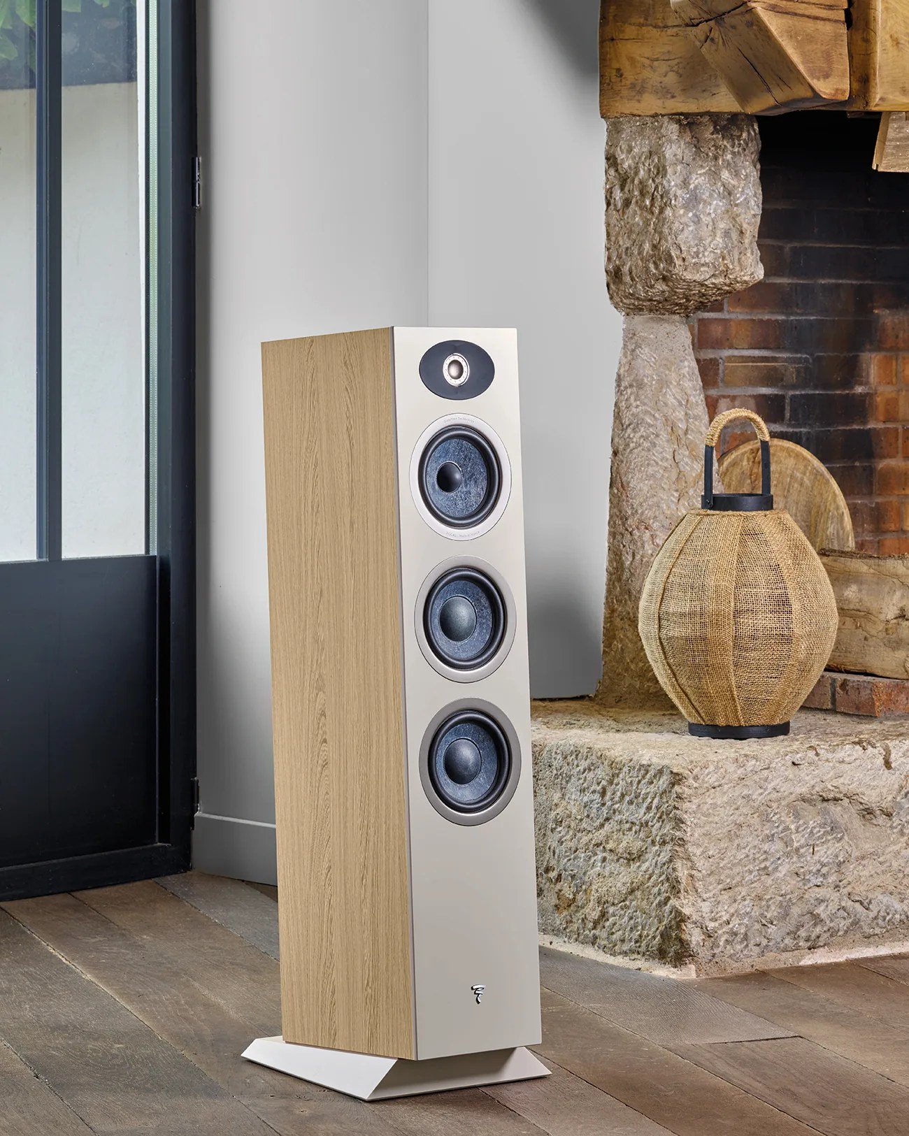 Tall floor-standing speaker with light wood sides and white front panel featuring three drivers, placed on a wooden floor near a stone fireplace.