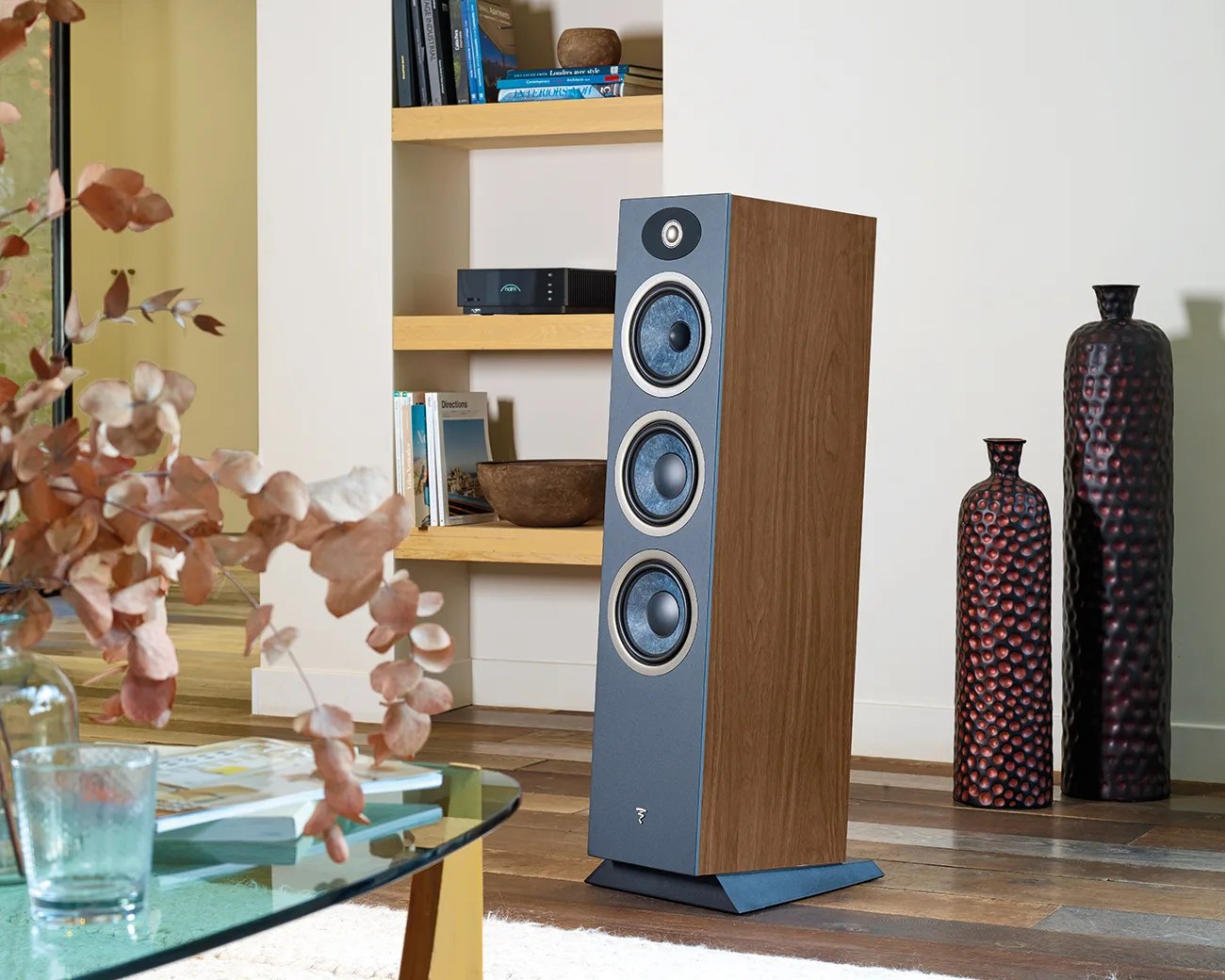 Floor-standing speaker with wood finish and three blue speaker cones in a modern living room setting.