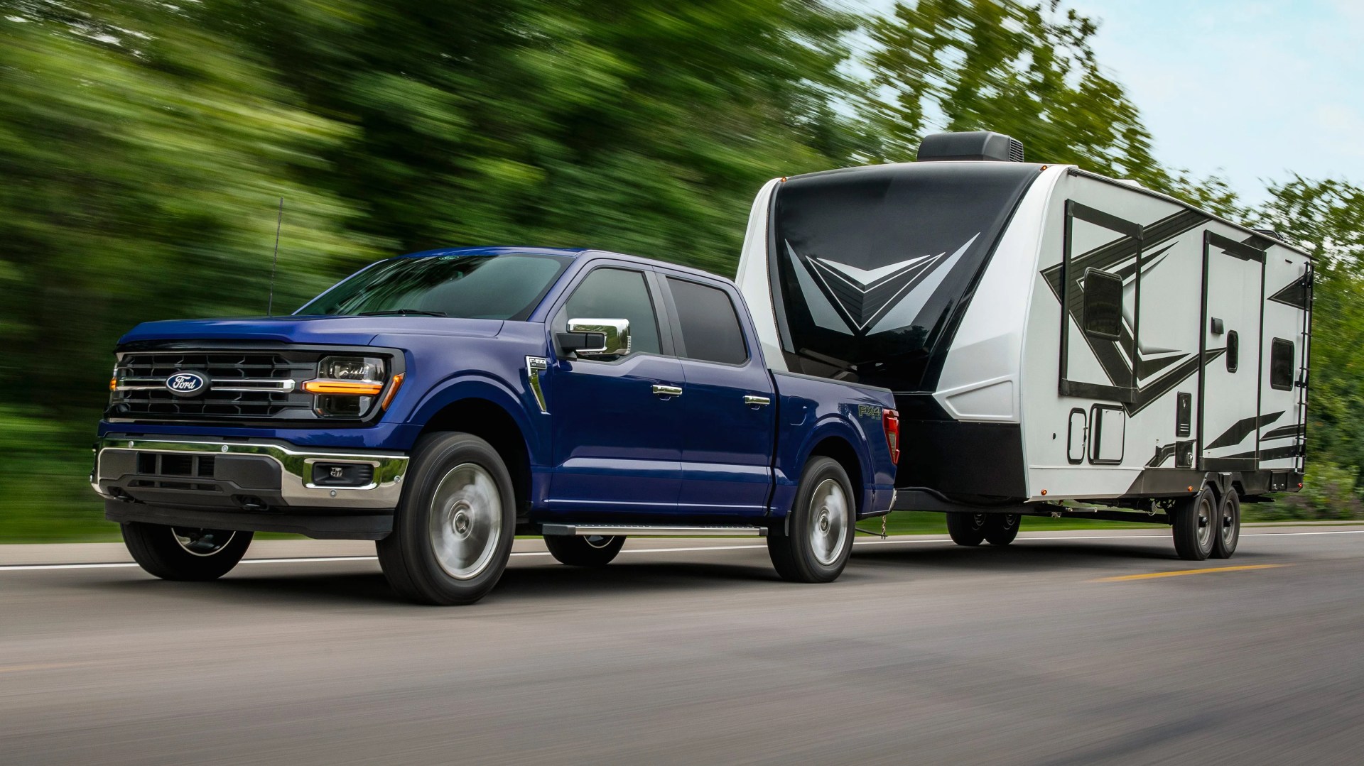 Blue Ford F-150 pickup truck towing a large white and black travel trailer on a road.