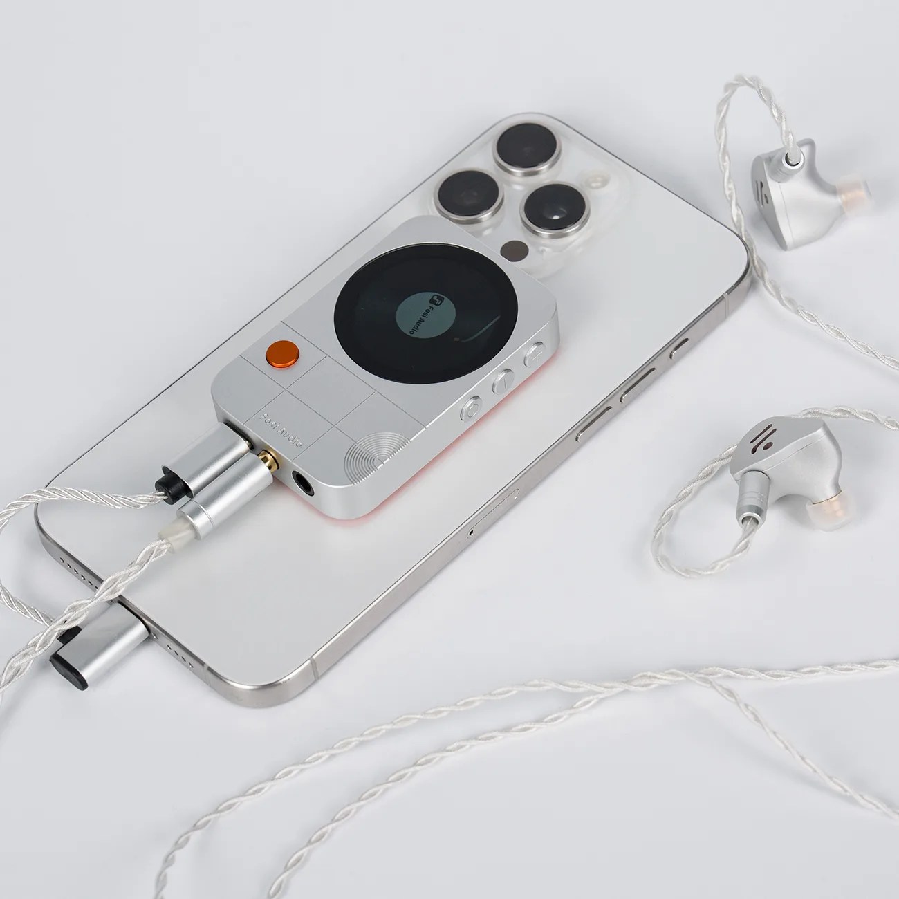 Silver portable music player with a round display and orange button connected to silver earphones, placed on a white smartphone.