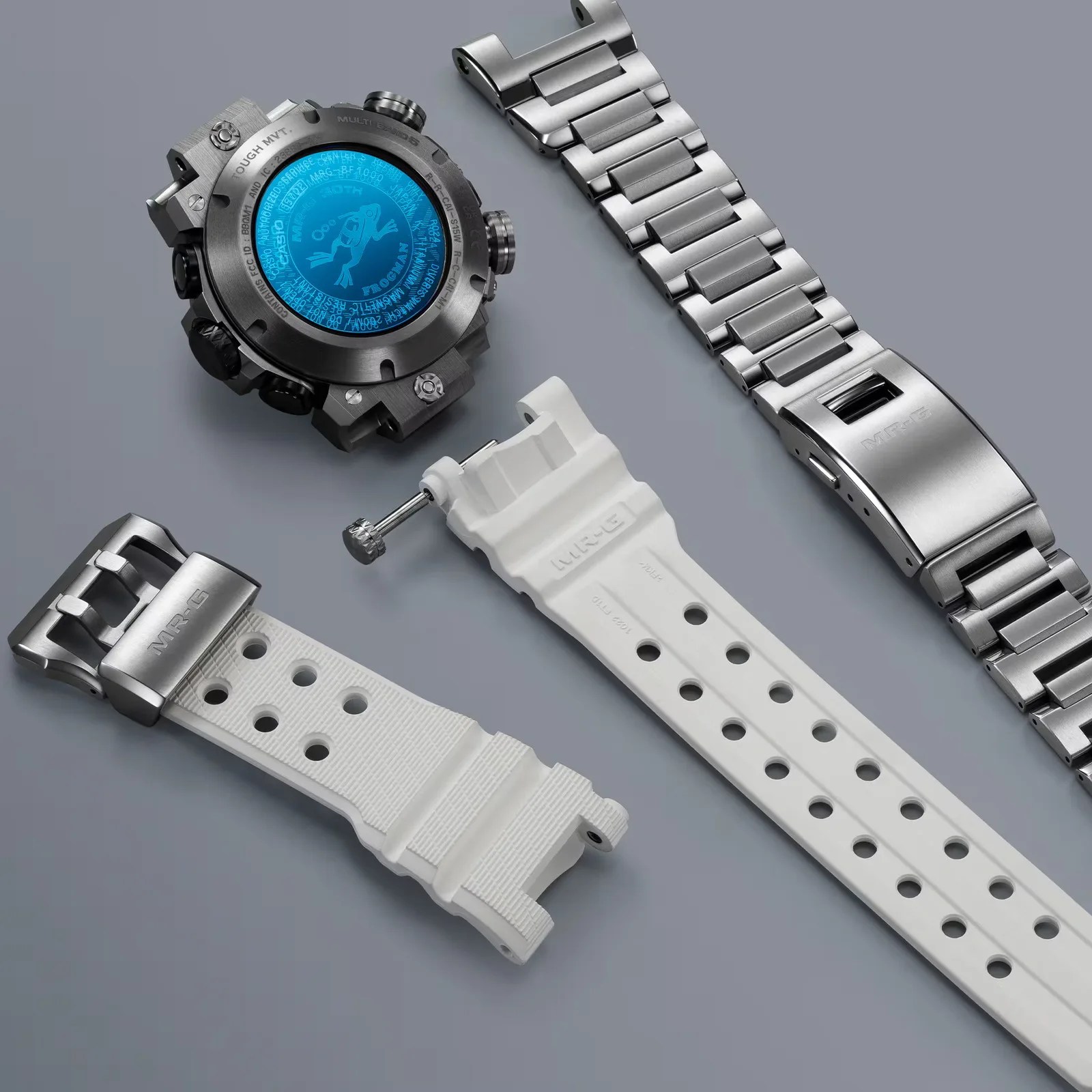 Disassembled Casio MR-G Frogman watch with titanium case, blue back cover, white resin strap, and metal bracelet on gray surface.