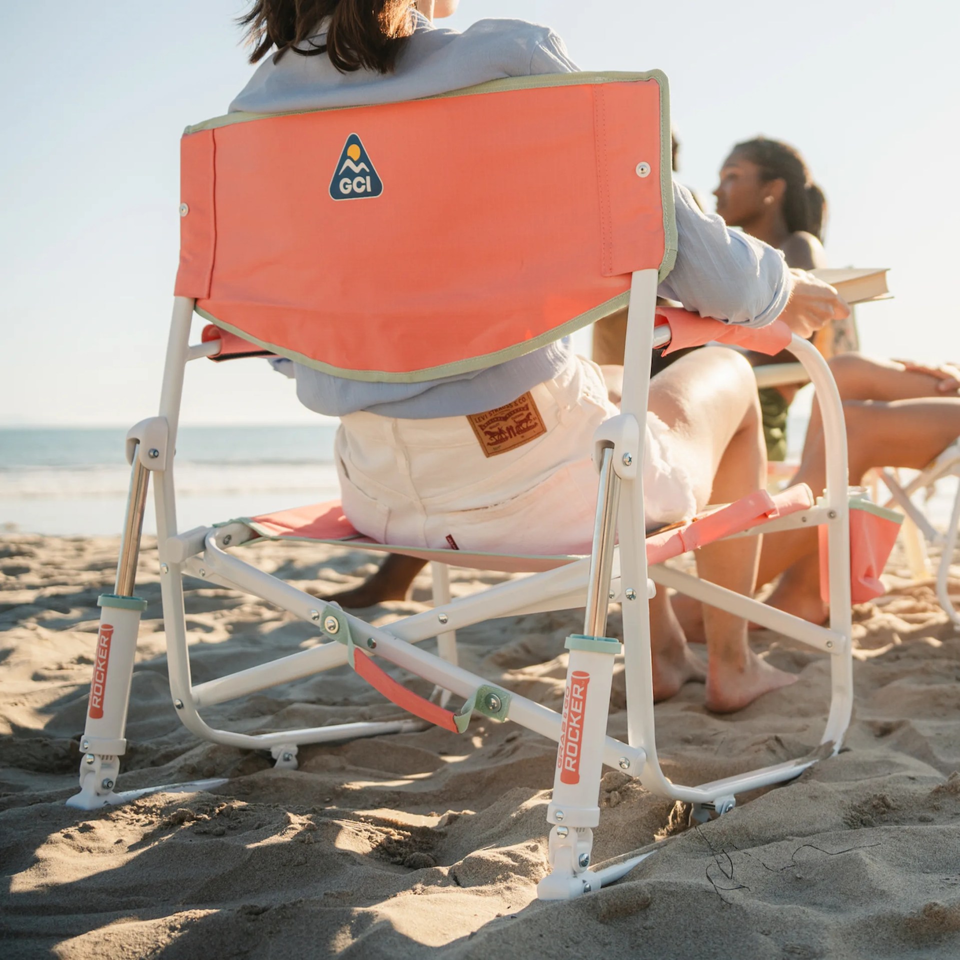 Orange GCI outdoor rocking chair with white frame on sandy beach with two people sitting in it.
