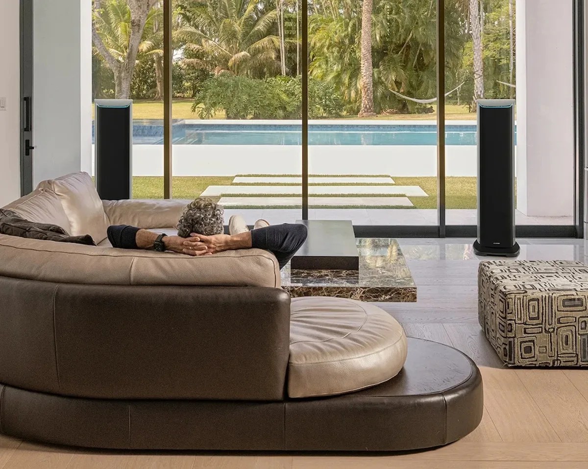 Man reclining on a curved beige and brown leather sofa facing a glass wall with a pool and garden outside.