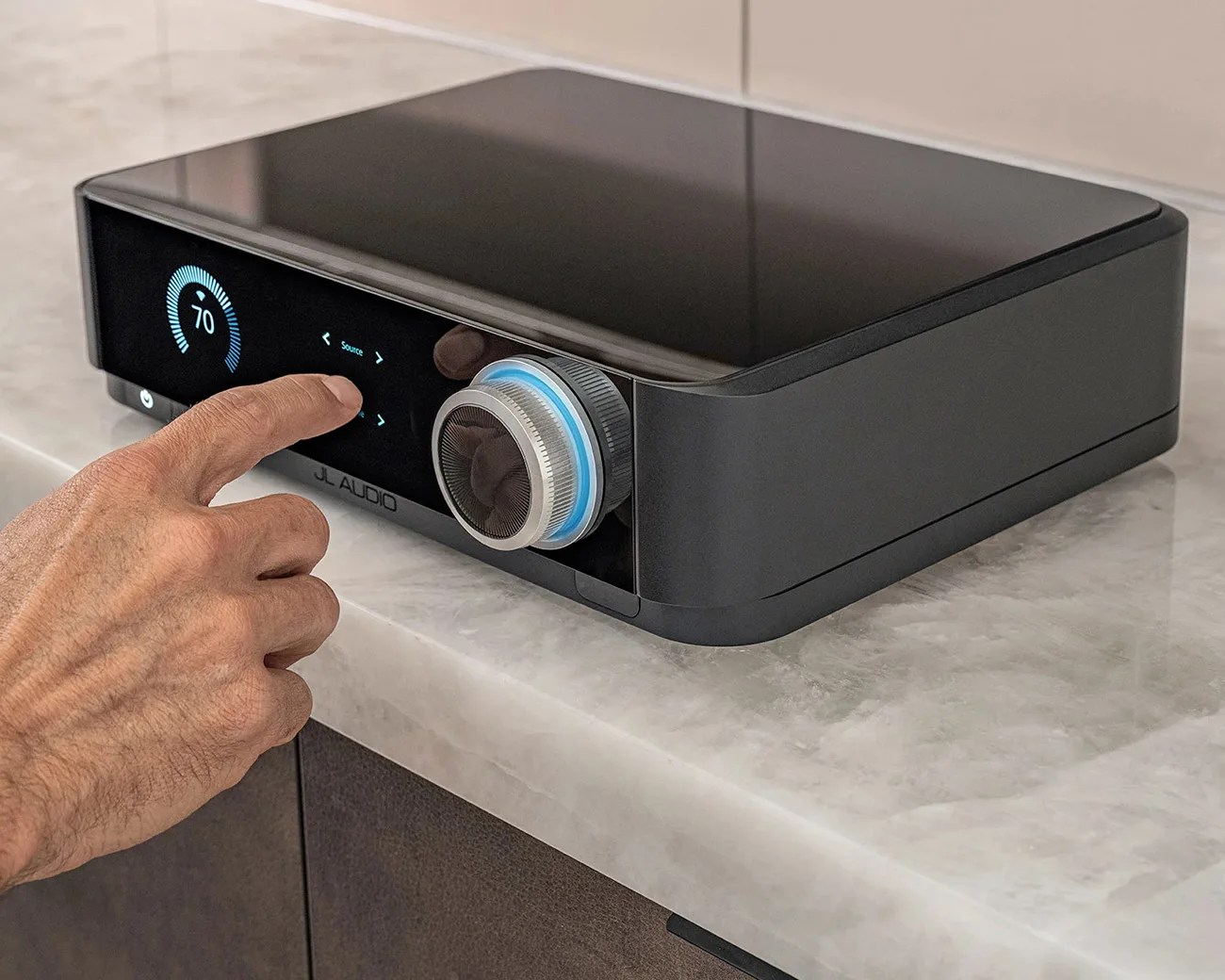 Black JL Audio device with digital display and large silver volume knob on a marble countertop, finger touching the screen.