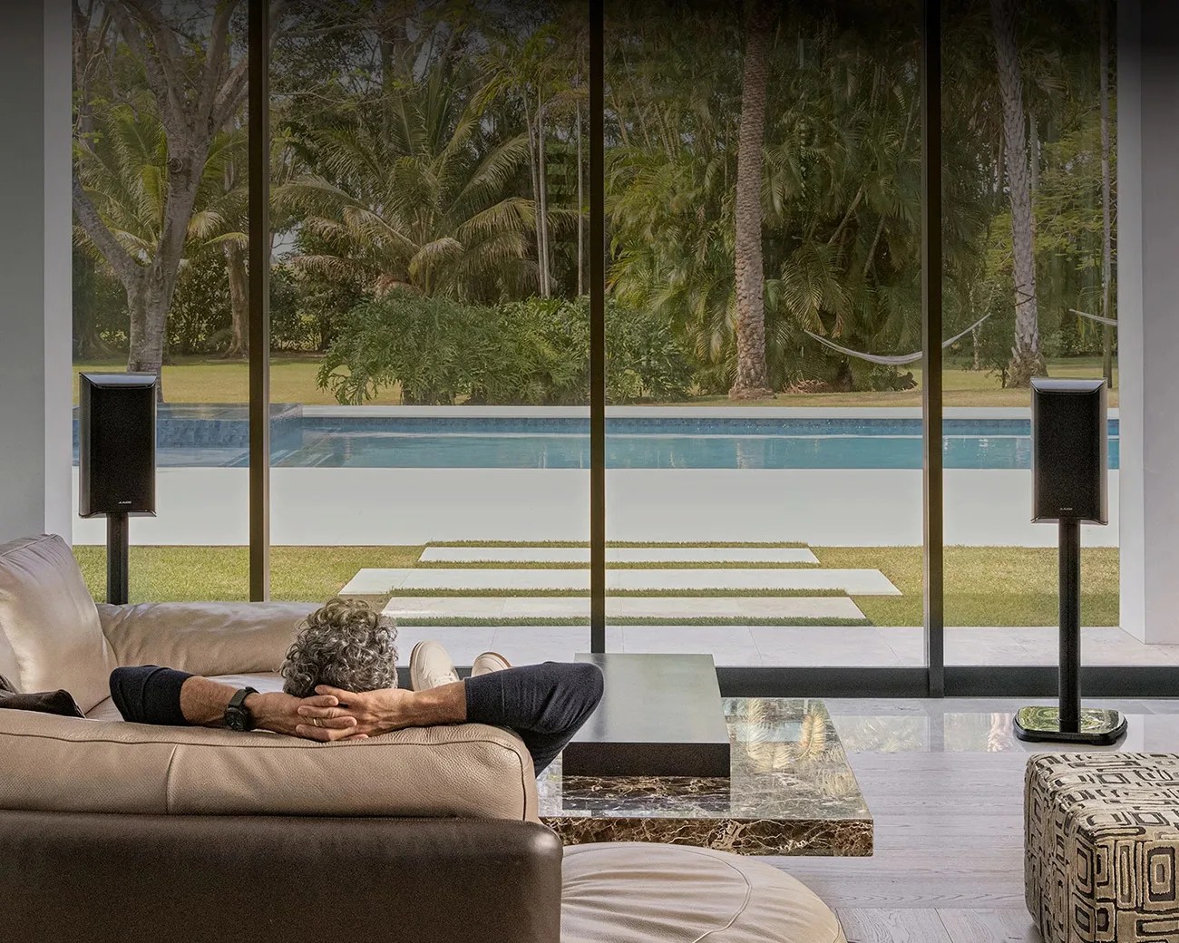 Person reclining on a beige leather sofa facing large glass windows overlooking a pool and tropical garden.