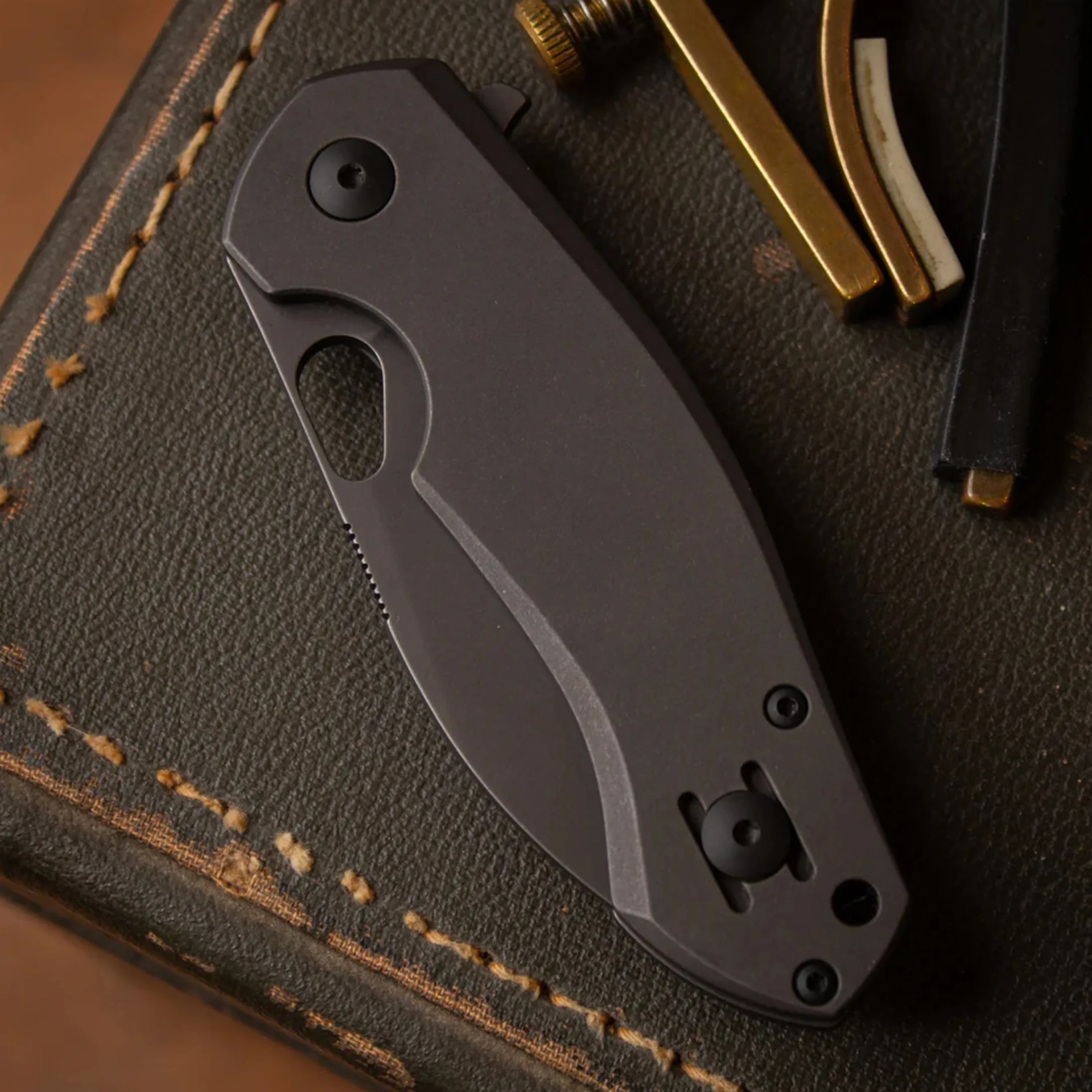 Closed matte black folding knife on a dark green leather surface with yellow stitching.