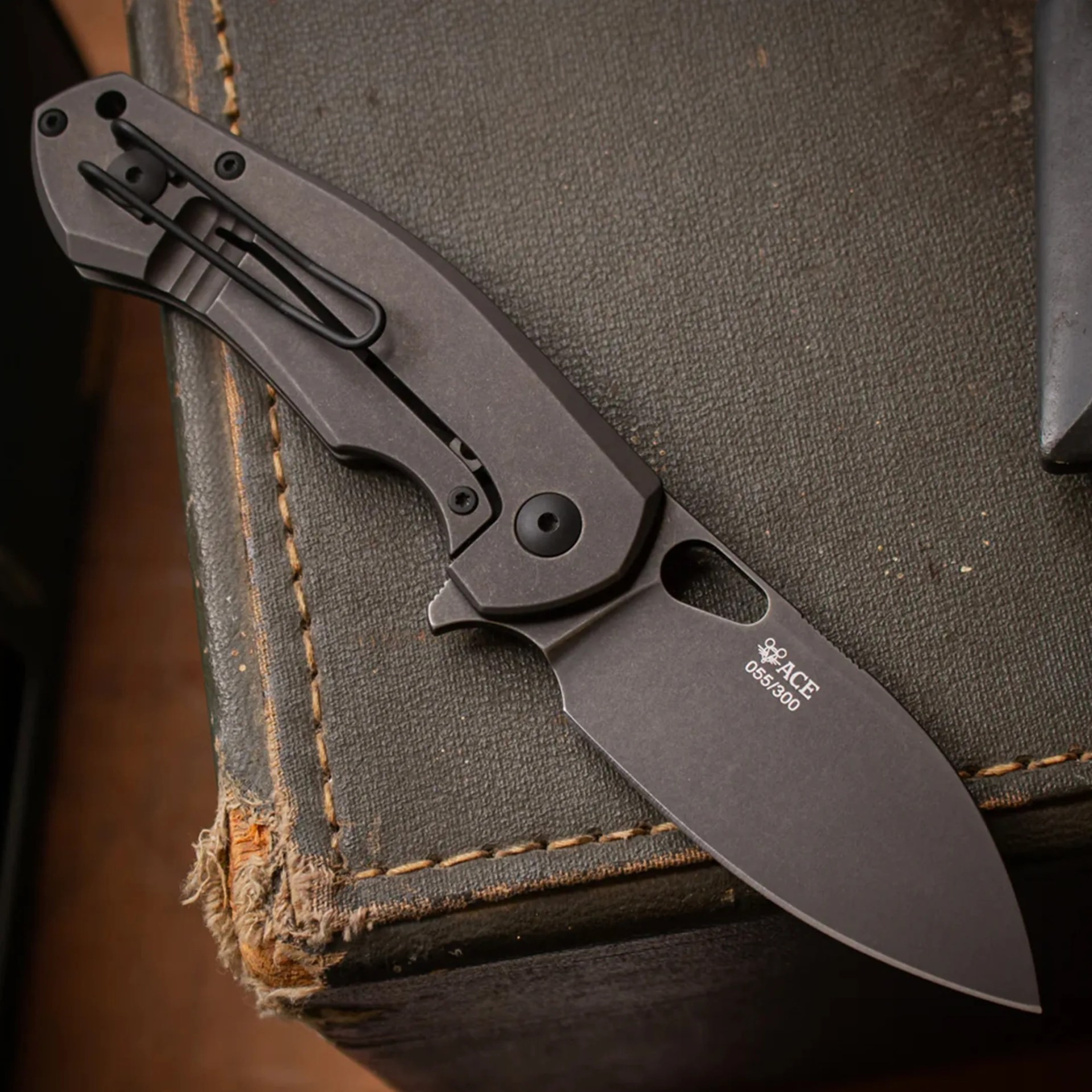 Black folding knife with clip and "ACE 055/300" text on blade resting on worn black fabric surface.