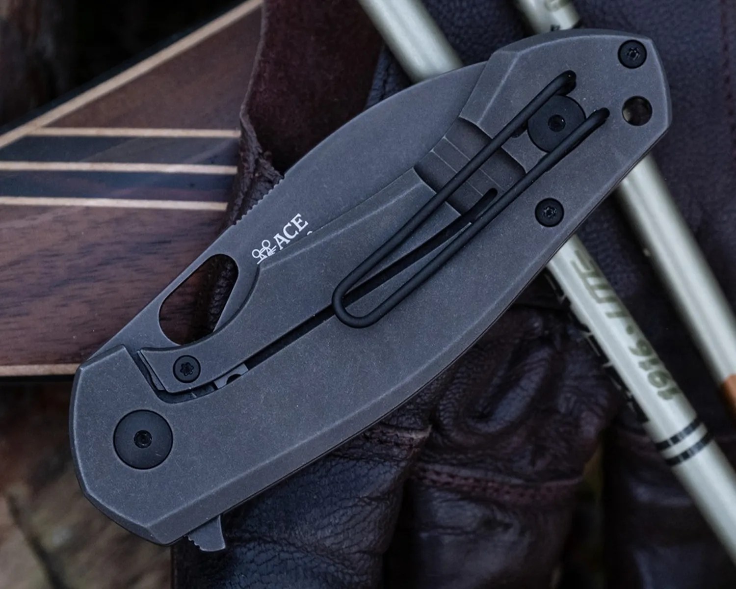 Folded dark gray metal pocket knife with black clip and "ACE" logo on a dark textured surface.