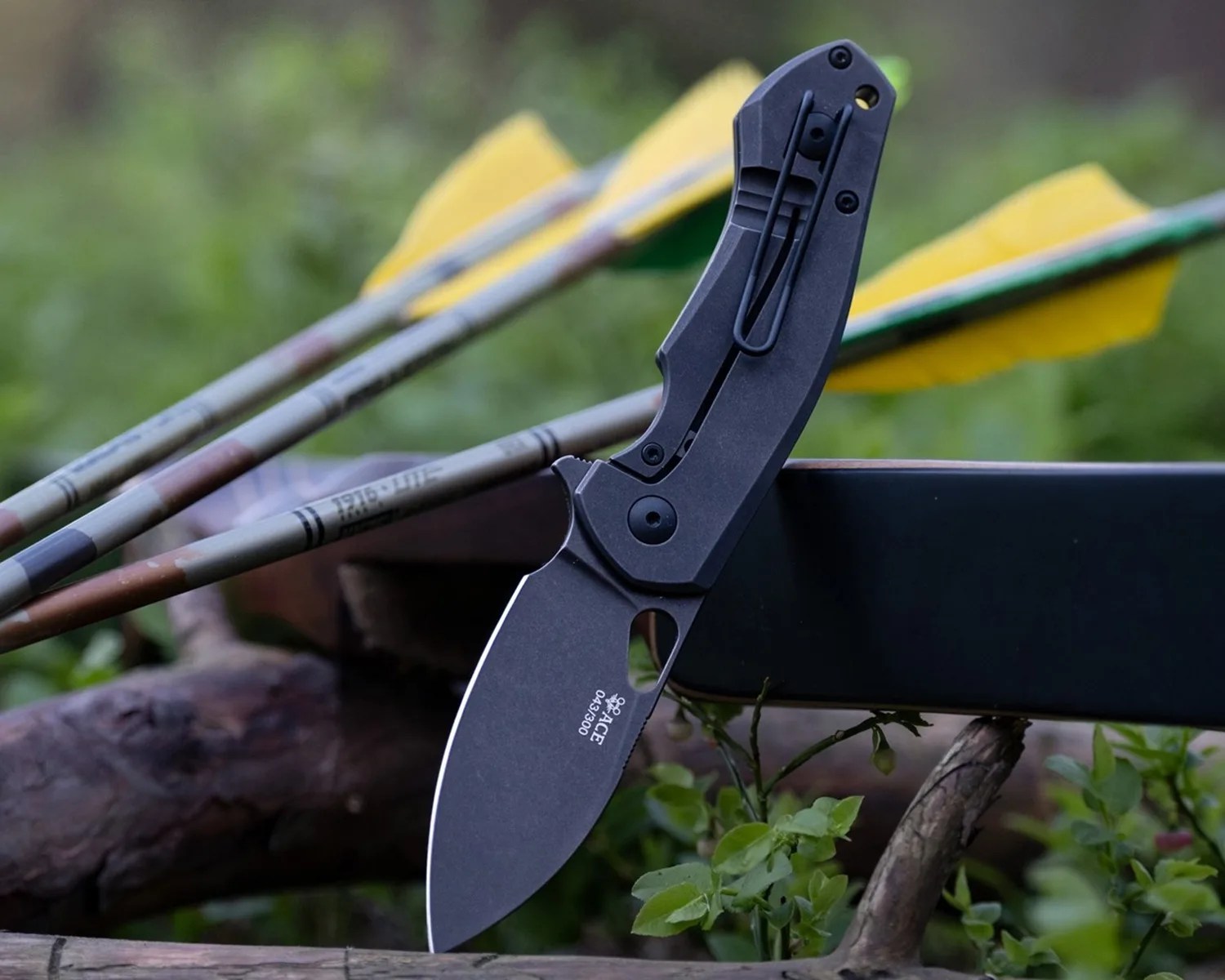 Black folding knife with clip and partially serrated blade resting on a black surface outdoors near arrows with yellow fletching.