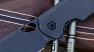 Close-up of a dark gray folding knife blade and handle with a thumb hole and pivot screw on a wooden surface.