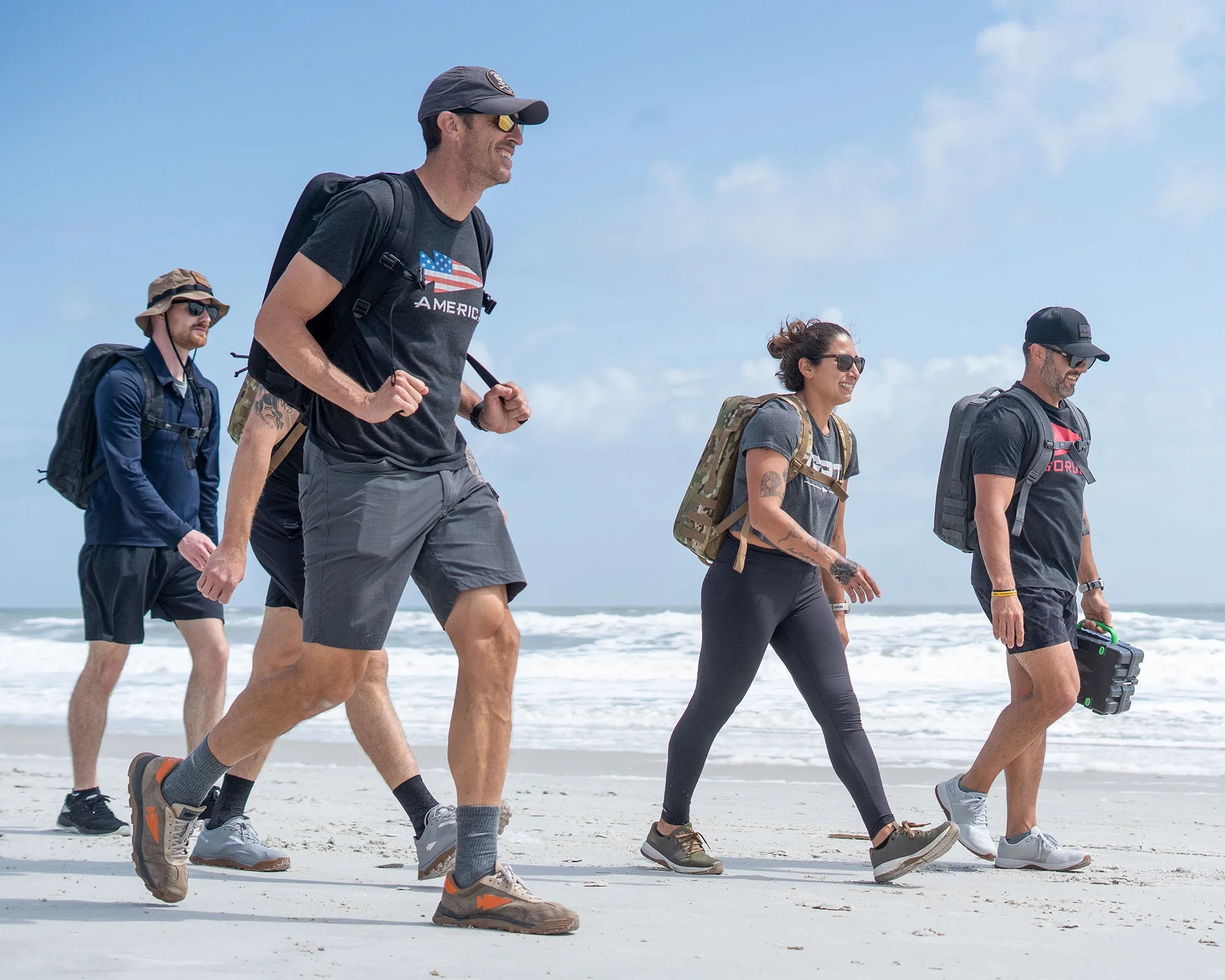 Group of four people walking on a sandy beach wearing backpacks, sunglasses, and casual outdoor clothing.
