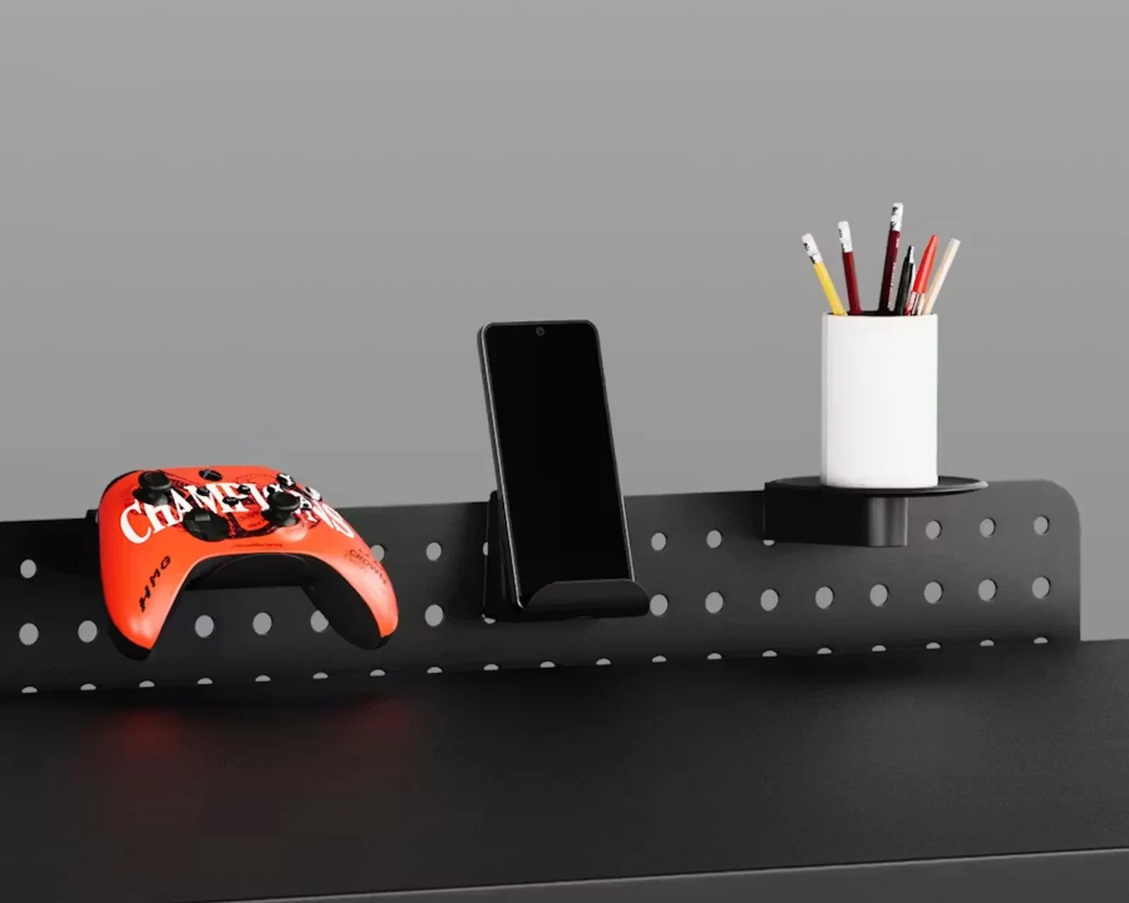 Black perforated metal wall organizer holding an orange game controller, a smartphone, and a white cup with pencils.