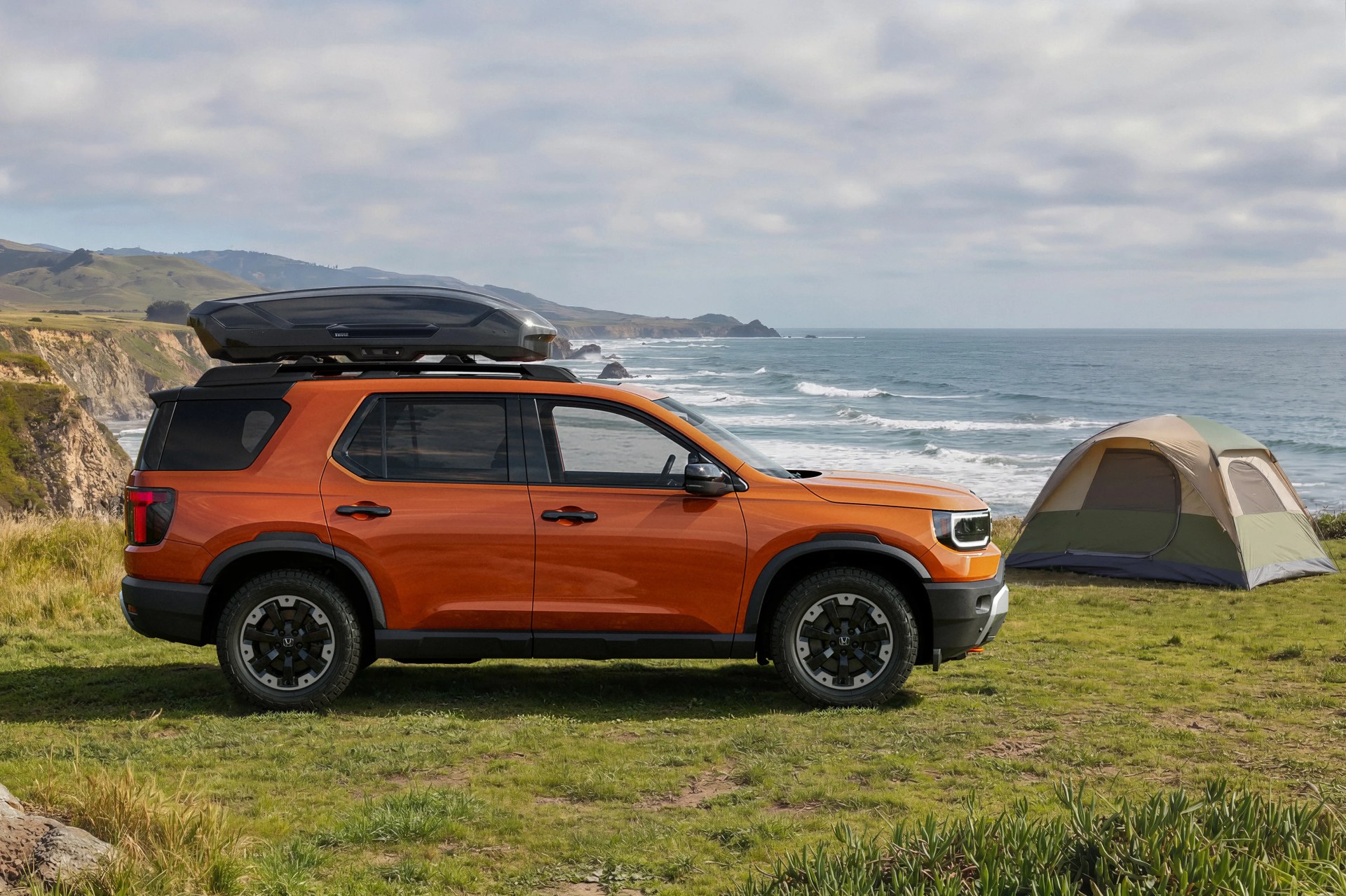 Orange SUV with black roof cargo box parked on grassy cliffside near a green and beige camping tent by the ocean.