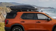 Orange SUV with black roof cargo box mounted on roof racks, parked near a coastal cliffside.