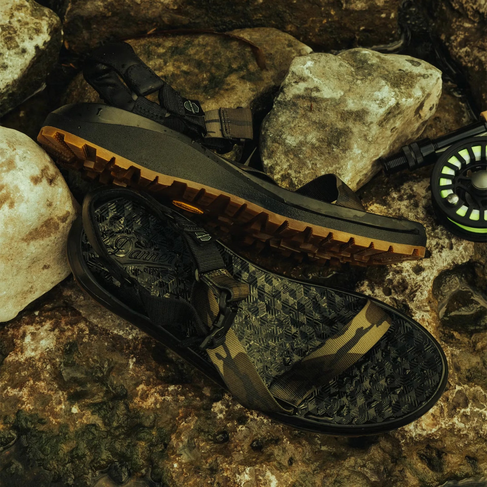 Pair of black and brown outdoor sandals with rugged soles on rocky ground near a fishing reel.