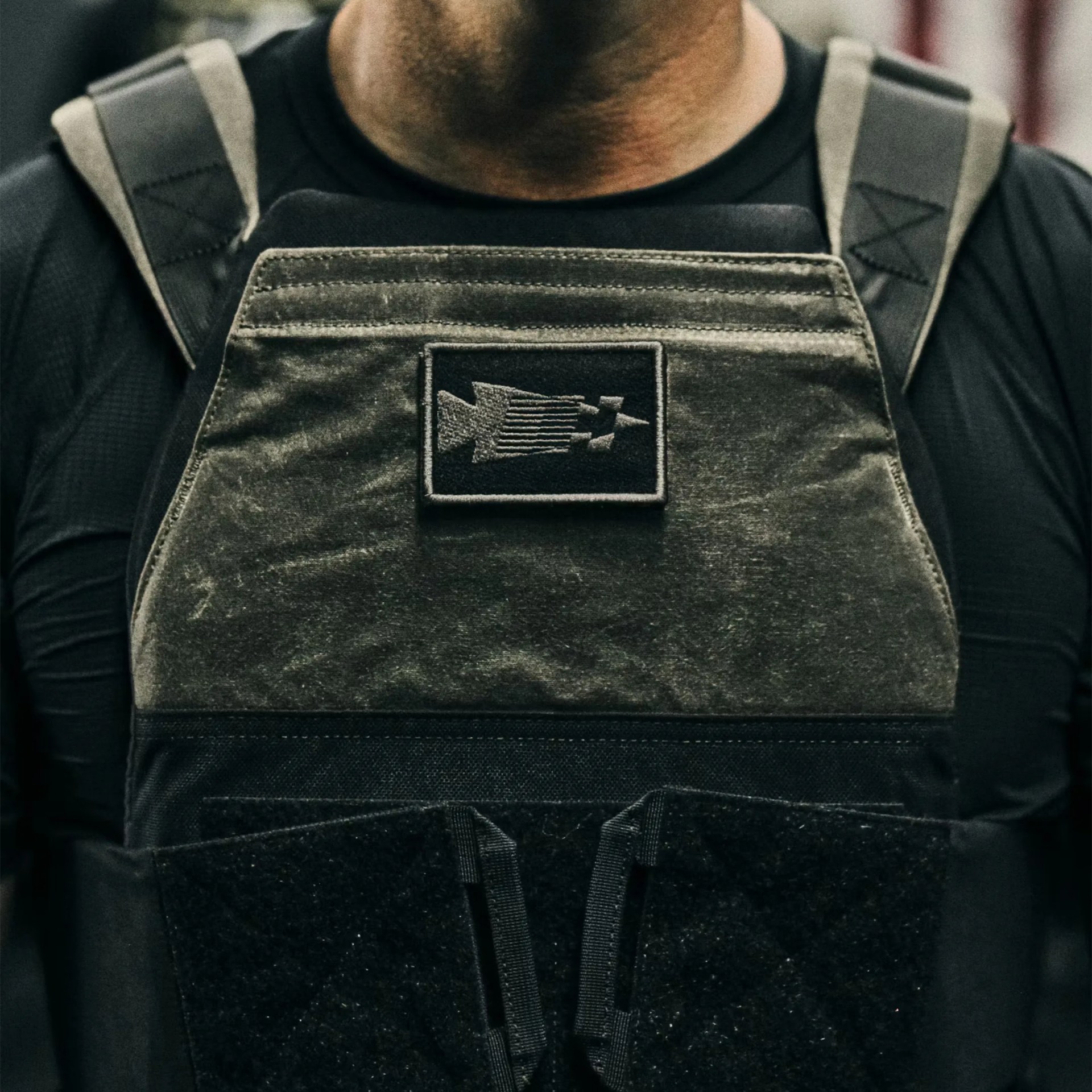 Close-up of a person wearing an olive green tactical vest with black straps and a patch featuring a stylized arrow design.