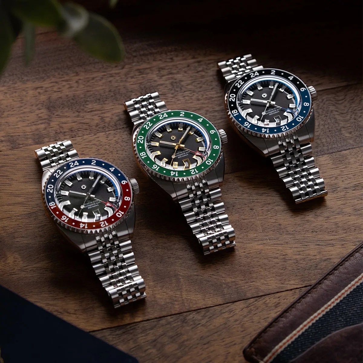 Three stainless steel wristwatches with multi-link bracelets and black dials, featuring red-blue, green, and blue-black 24-hour bezels, on a wooden surface.