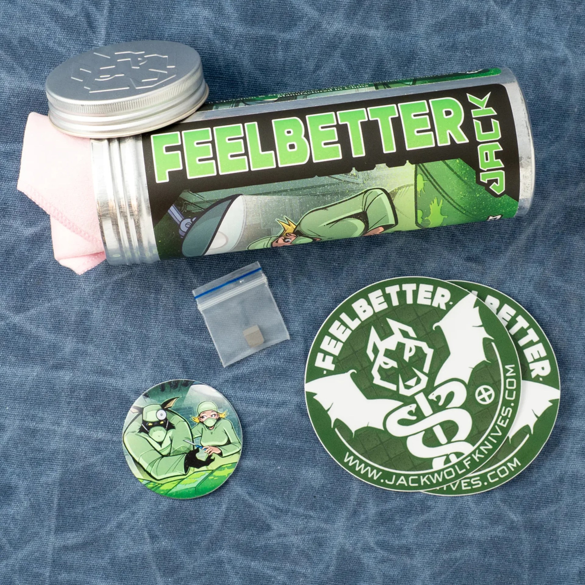 Silver metal container with a green "FEELBETTER JACK" label, a small plastic bag, and two circular stickers on blue fabric.