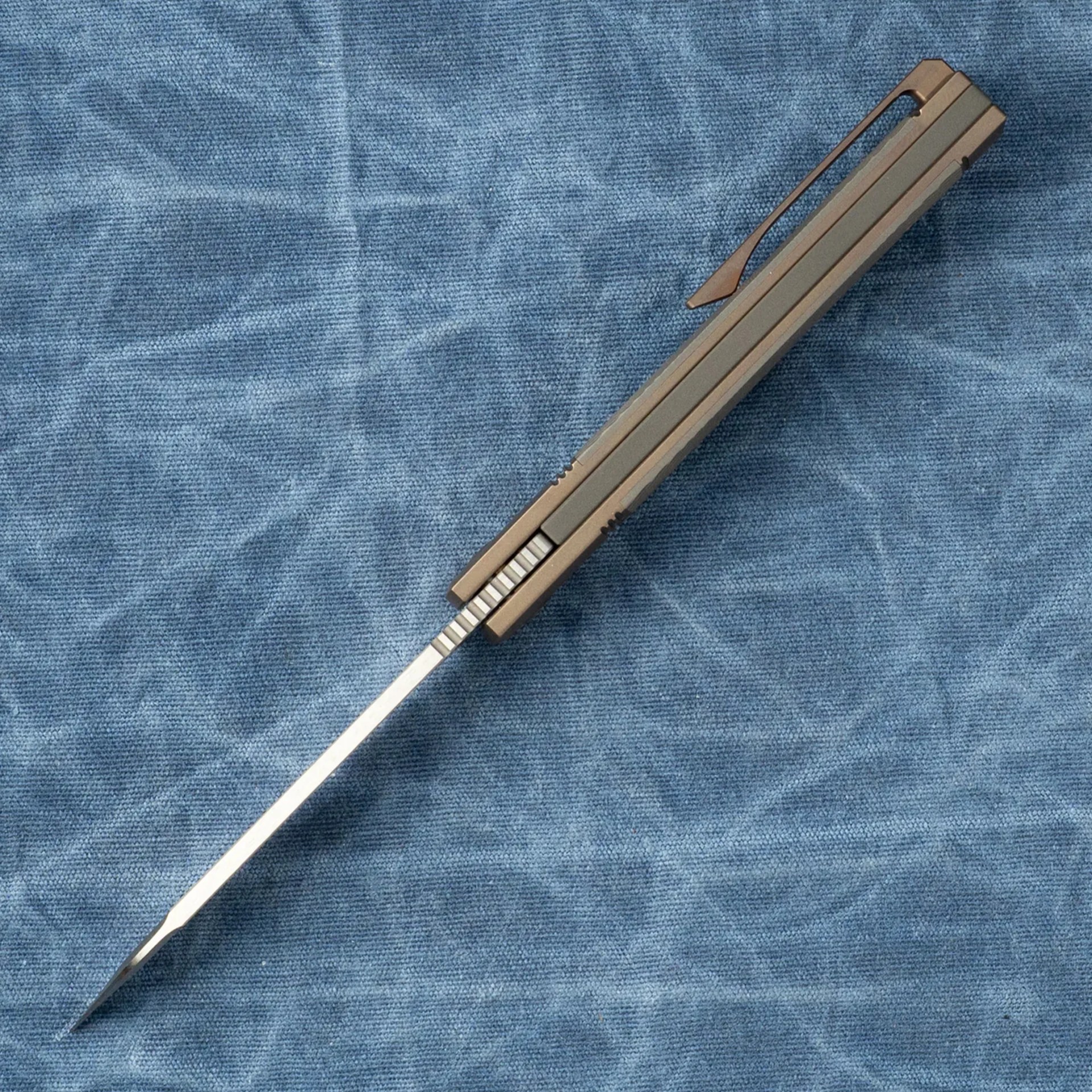 Slim metallic folding knife with a pocket clip on a blue textured fabric.