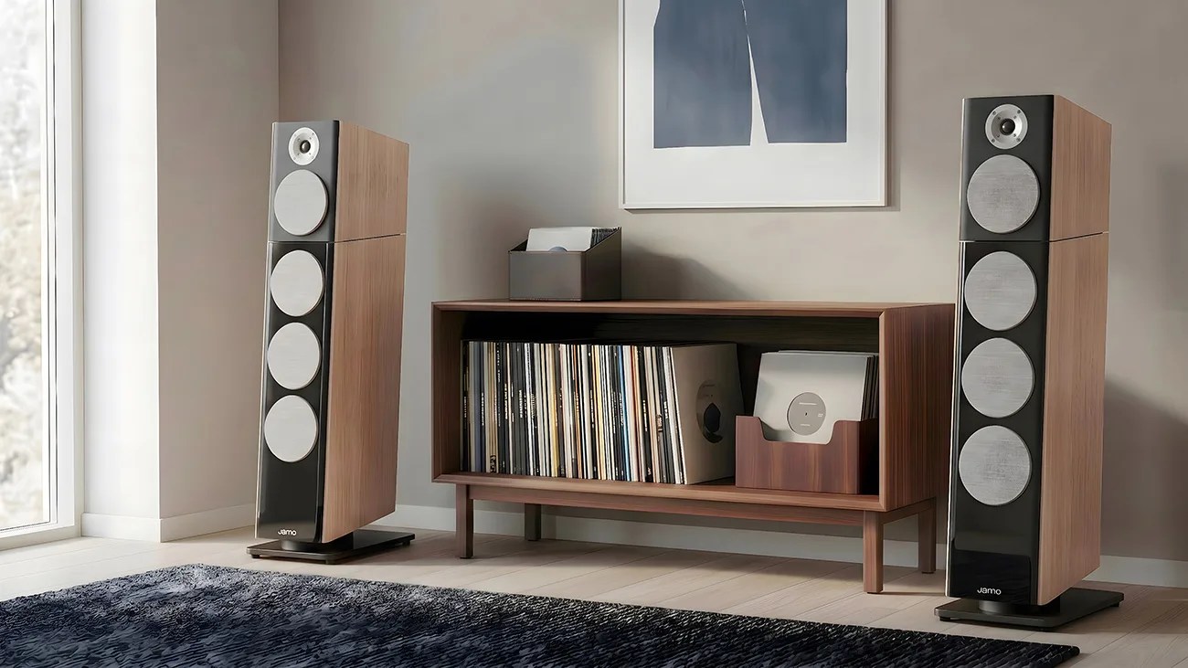 Pair of tall wooden floor-standing speakers with silver drivers flanking a wooden record shelf filled with vinyl records.
