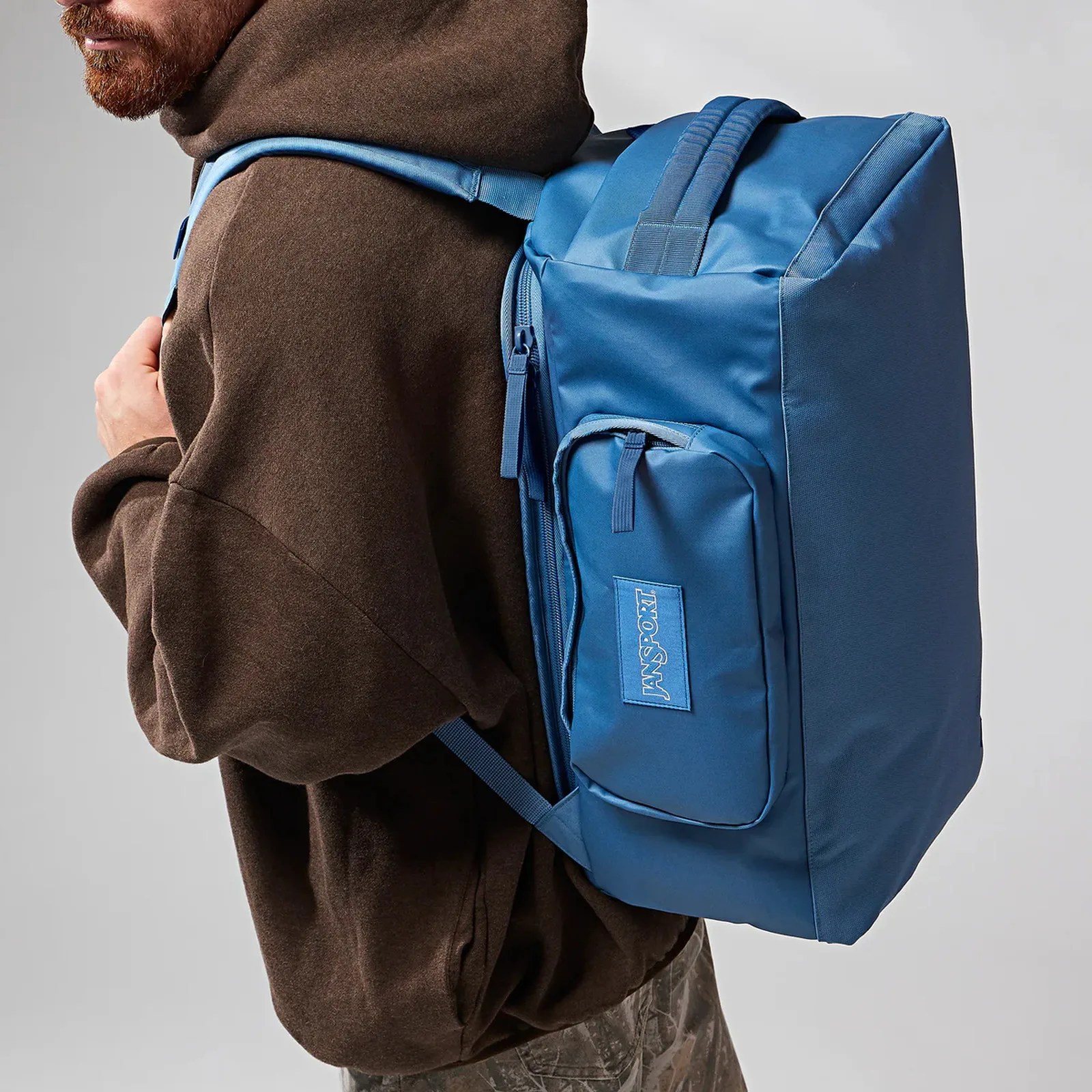 Blue Jansport backpack with front pocket worn by a person in a brown hoodie.