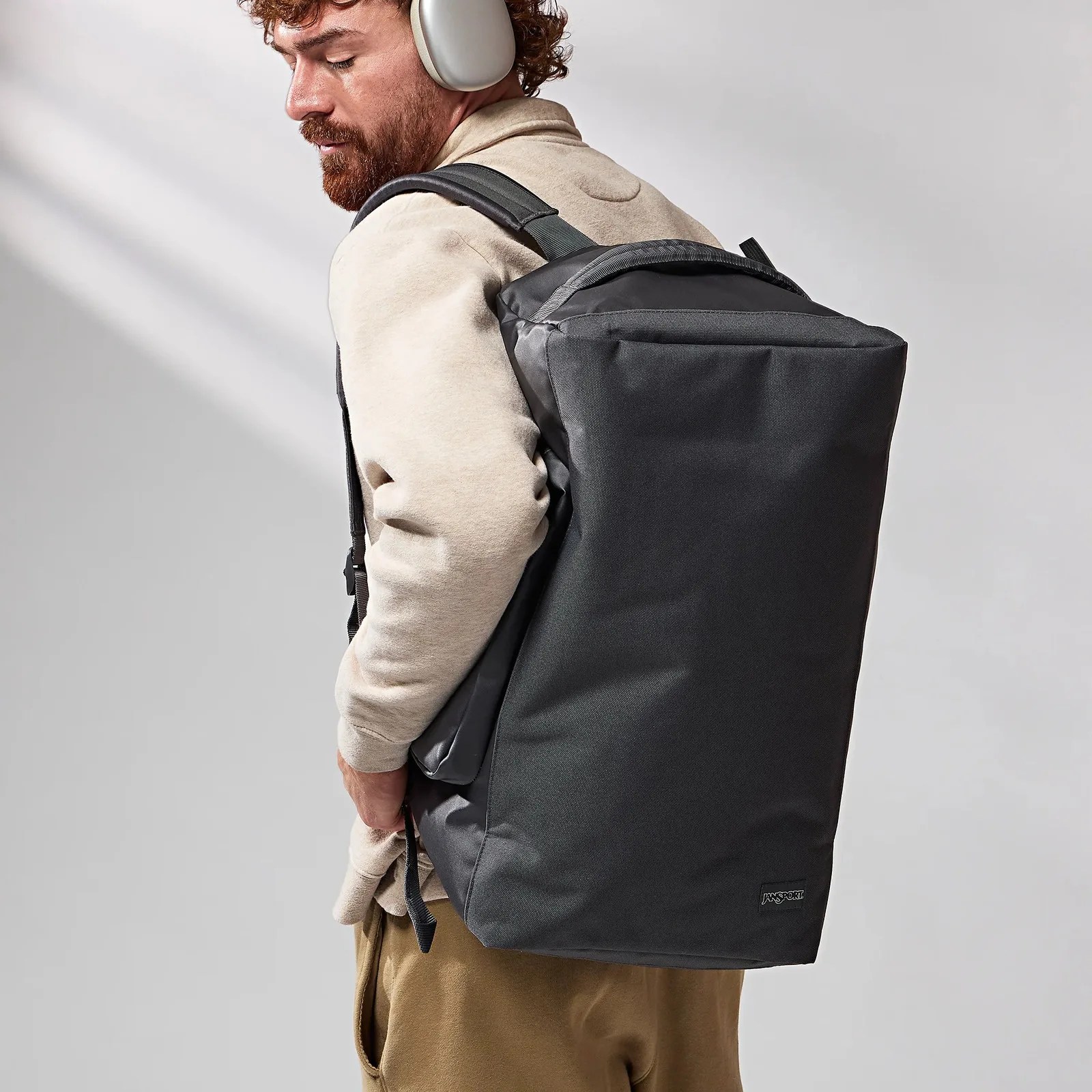 Black Jansport backpack with padded straps worn by a person in beige and brown clothing.
