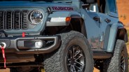 Close-up of a gray Jeep Rubicon front side showing headlight, grille, large off-road tire, and red tow strap on the bumper.